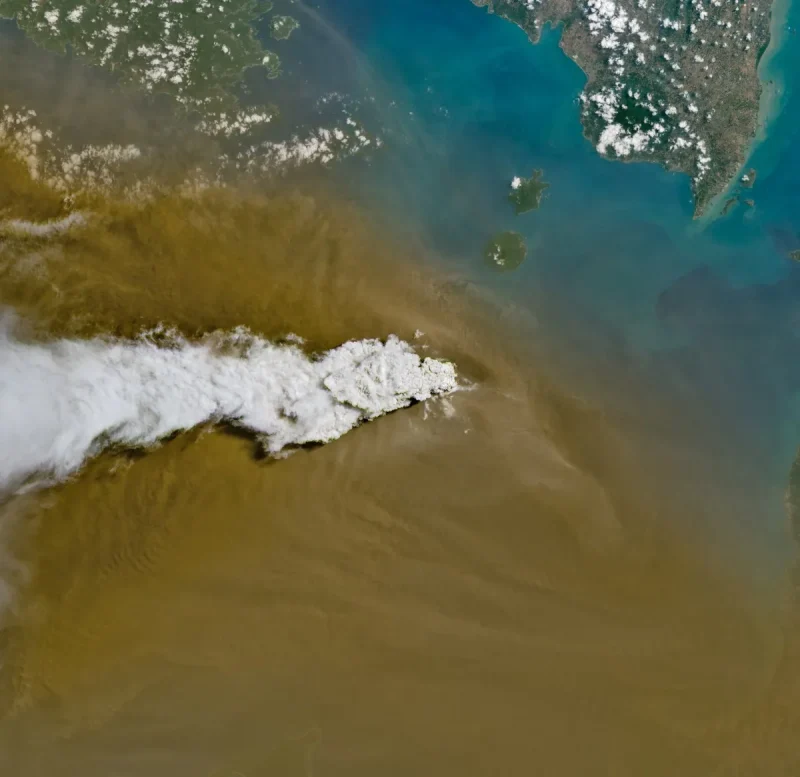 A white volcanic plume streams left over a tan ash cloud that fills most of the image. A small green island and blue water are visible in the upper right.