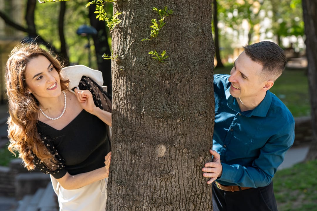 A couple looks at each other hidden behind a tree