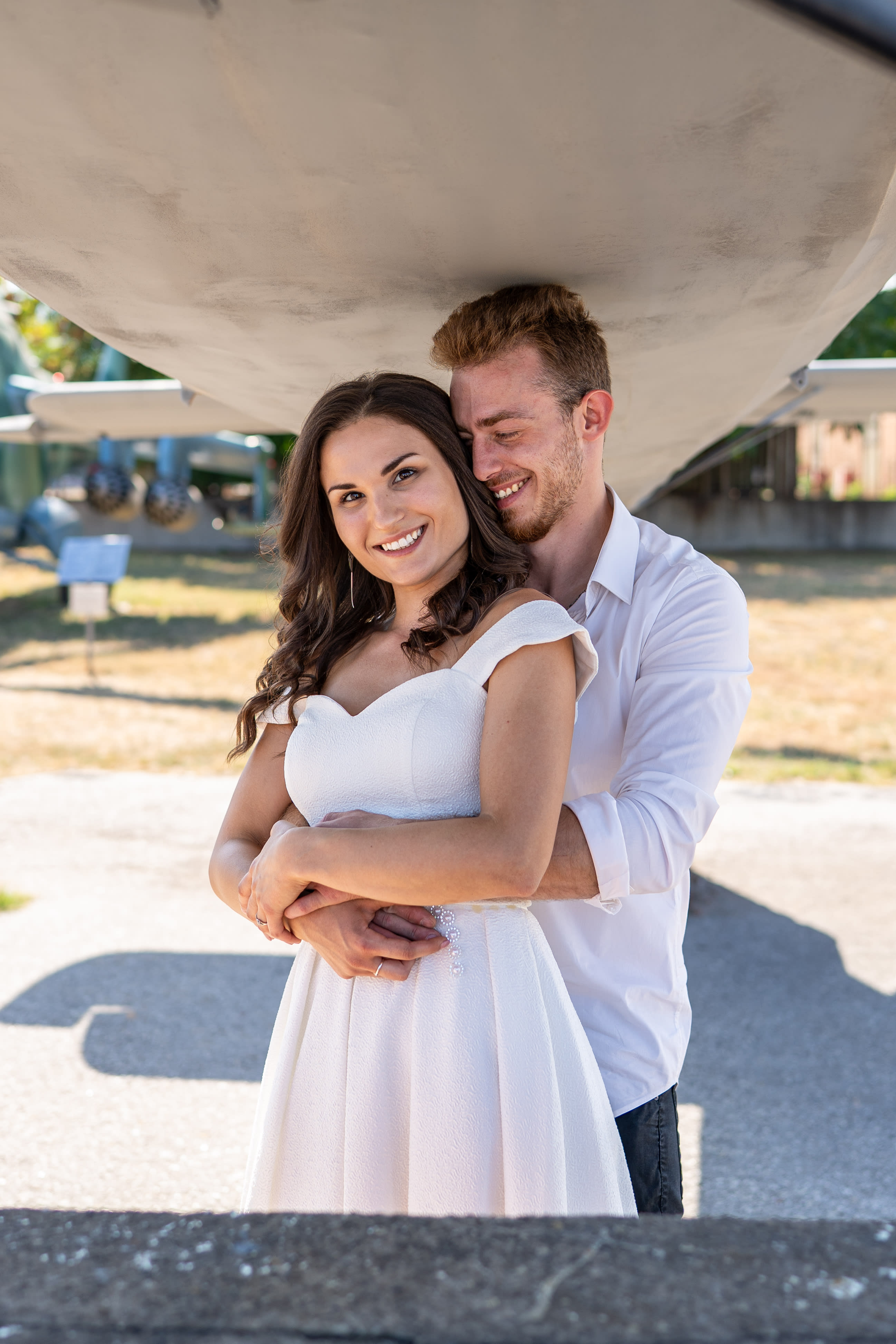 Couple hugging under the wing of an old airplane. The woman is in a white dress, and the man is in a white shirt.