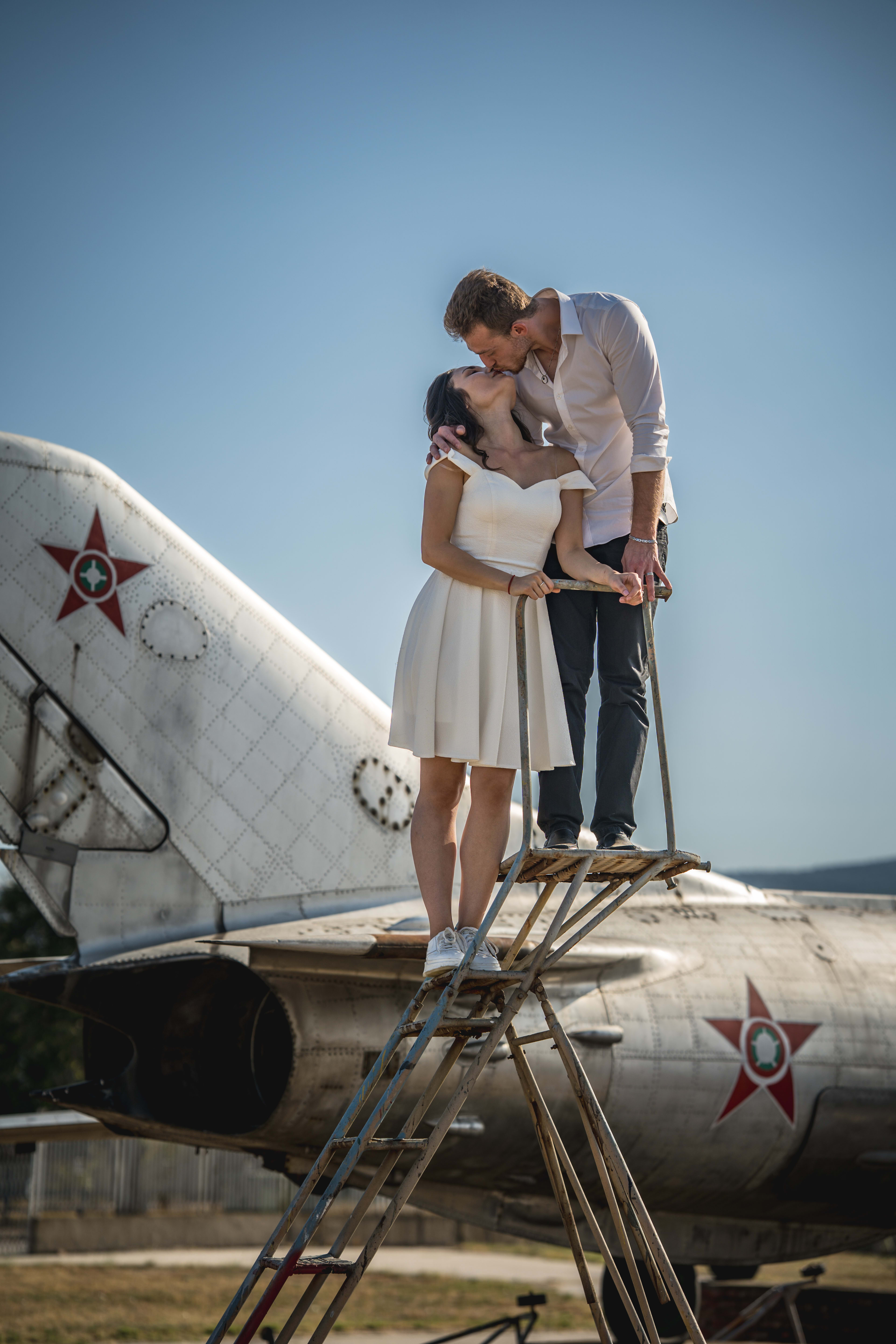Couple kissing on top of an old airplane. The woman is in a white dress, and the man is in a white shirt and jeans.