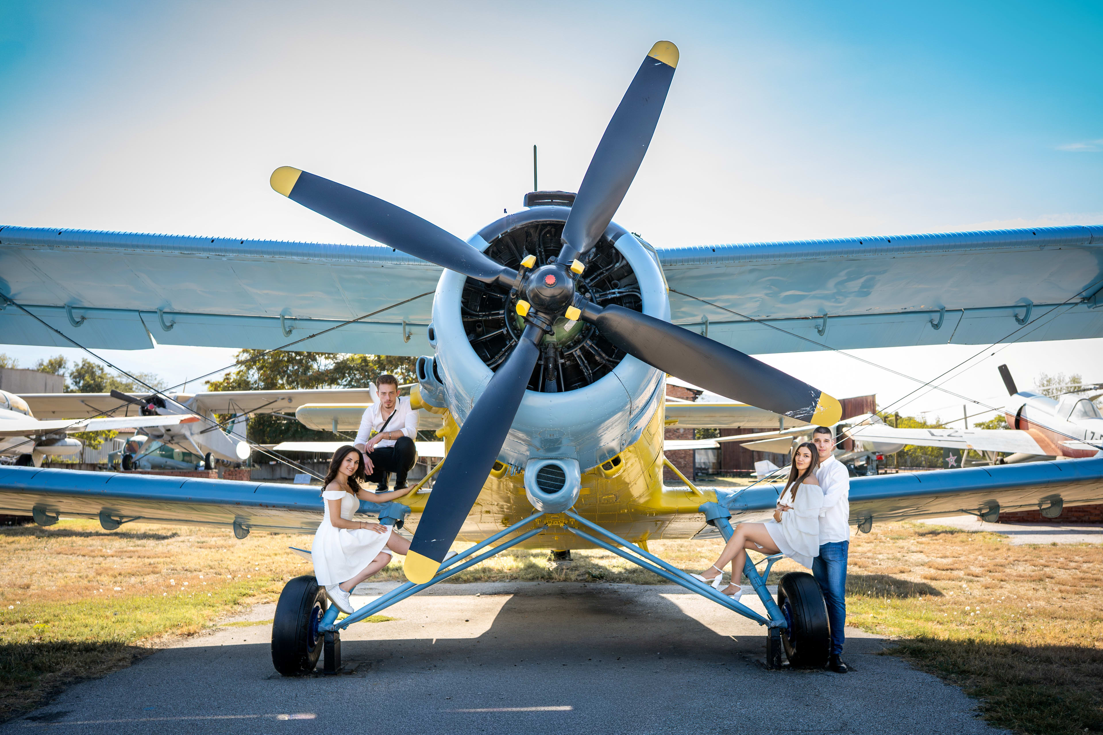 Two couples smiling and posing on the wings of an old airplane. The women are in white dresses, and the men are in white shirts and jeans.
