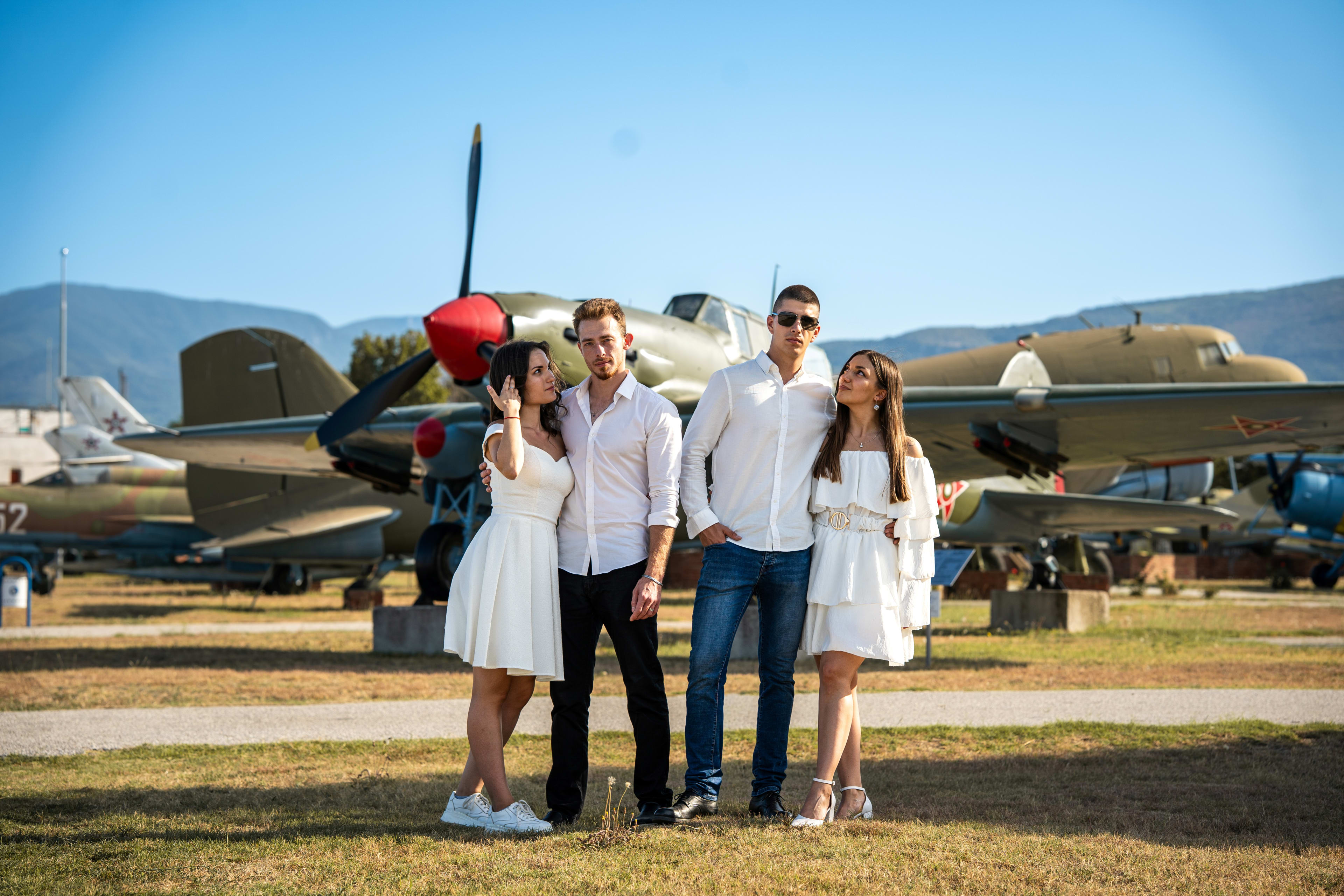 Two couples smiling and posing in a field of old airplanes. The women are in white dresses, and the men are in white shirts and jeans.