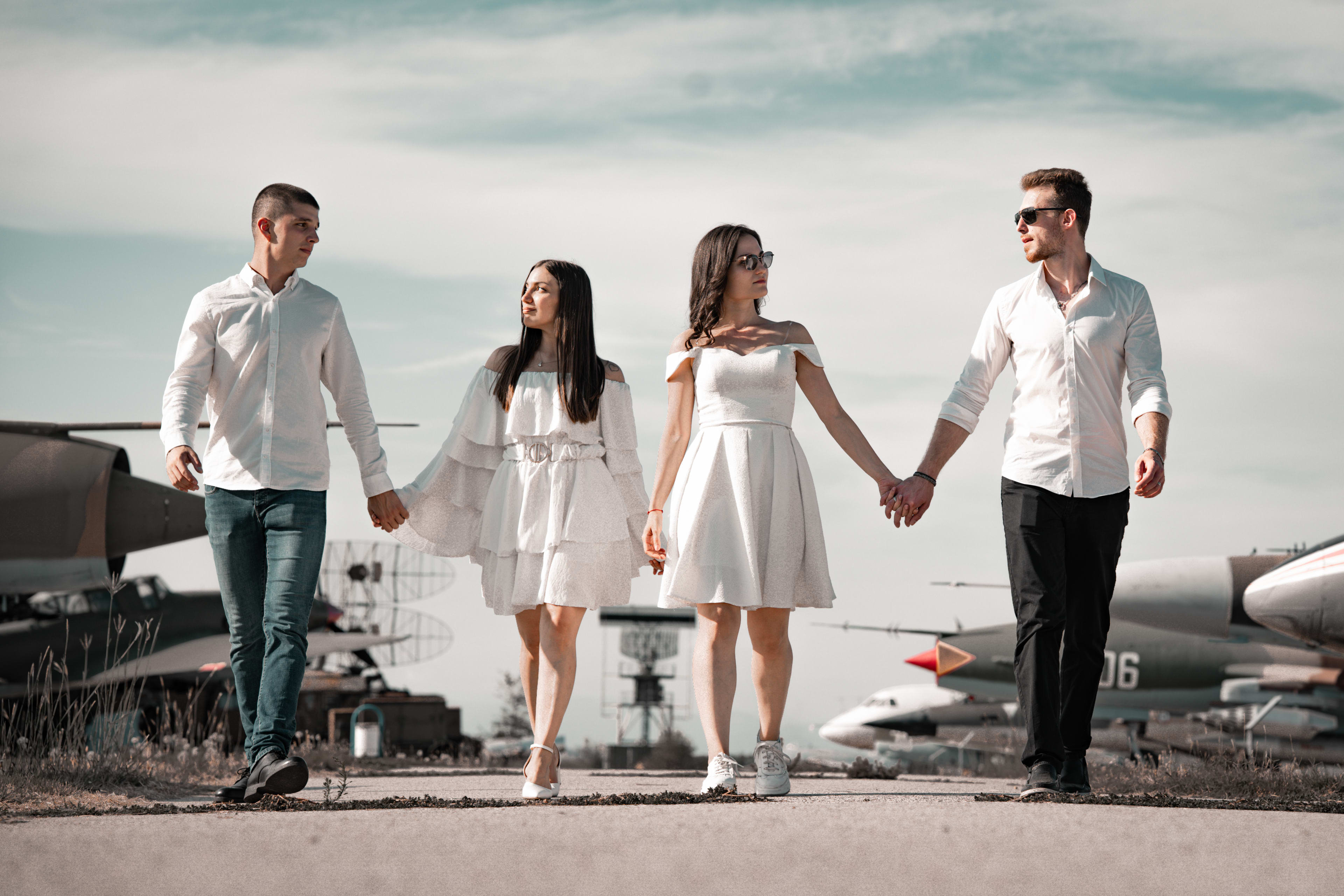 Two couples walking hand in hand in a field of old airplanes. The women are in white dresses, and the men are in white shirts and jeans.