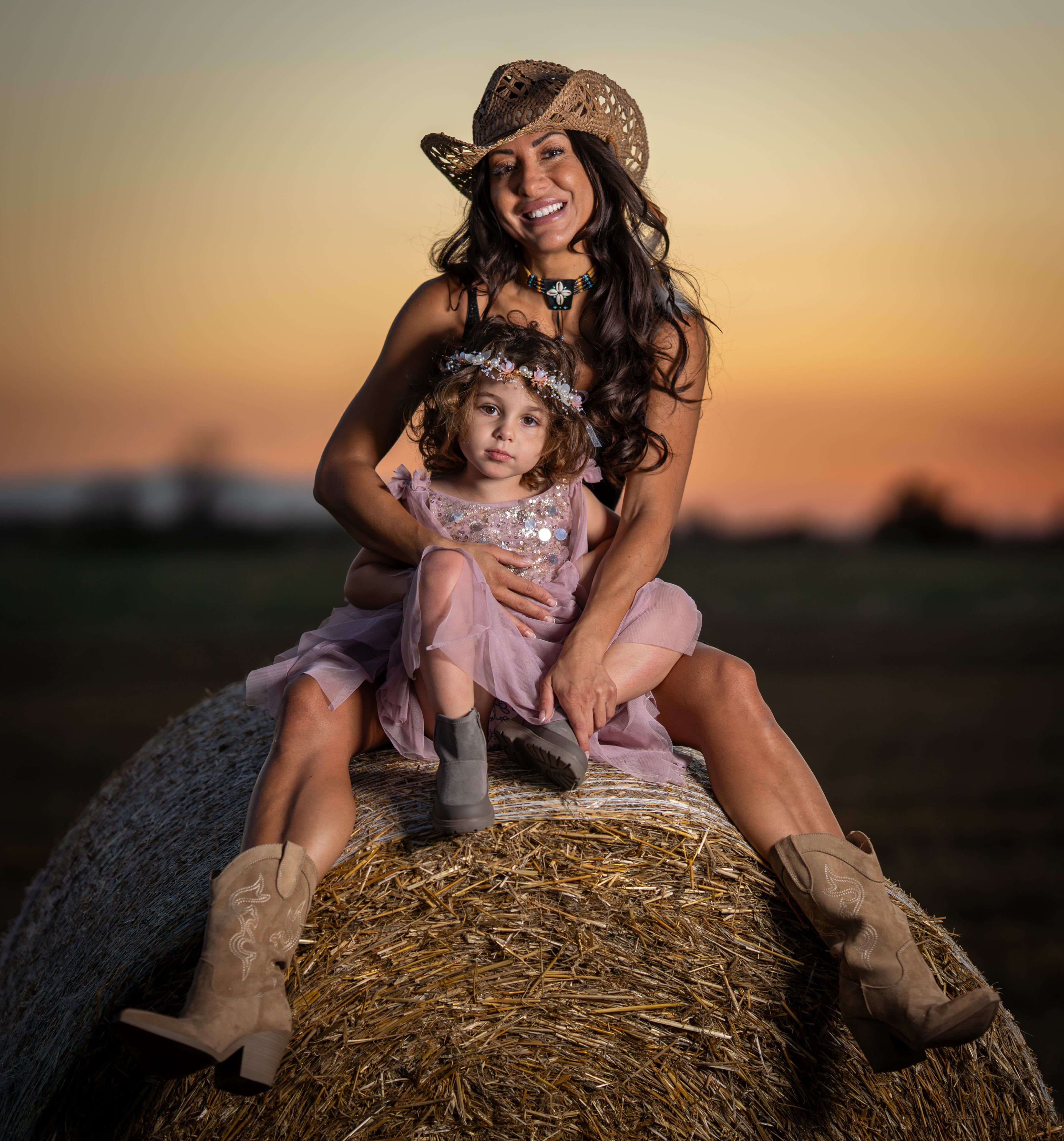 A cowgirl and her child sit on a straw bale