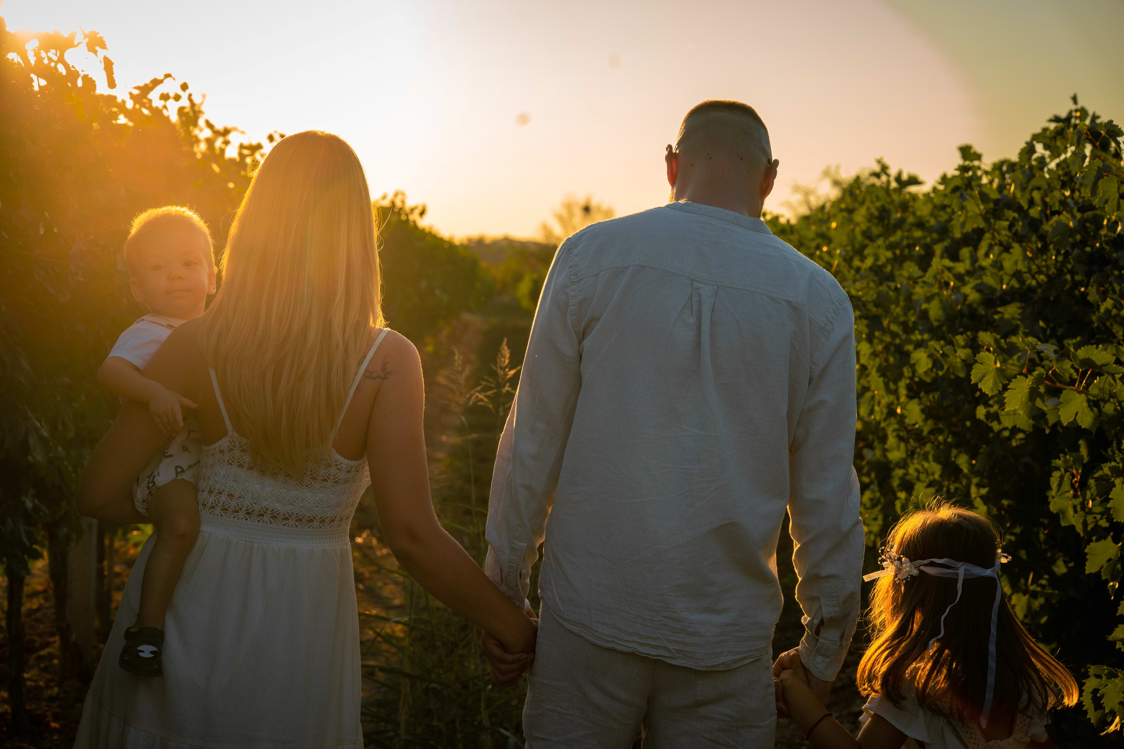 Mother, father, little girl, and little boy walking toward the sunset among the vineyards. Everyone is dressed in white and holding hands.