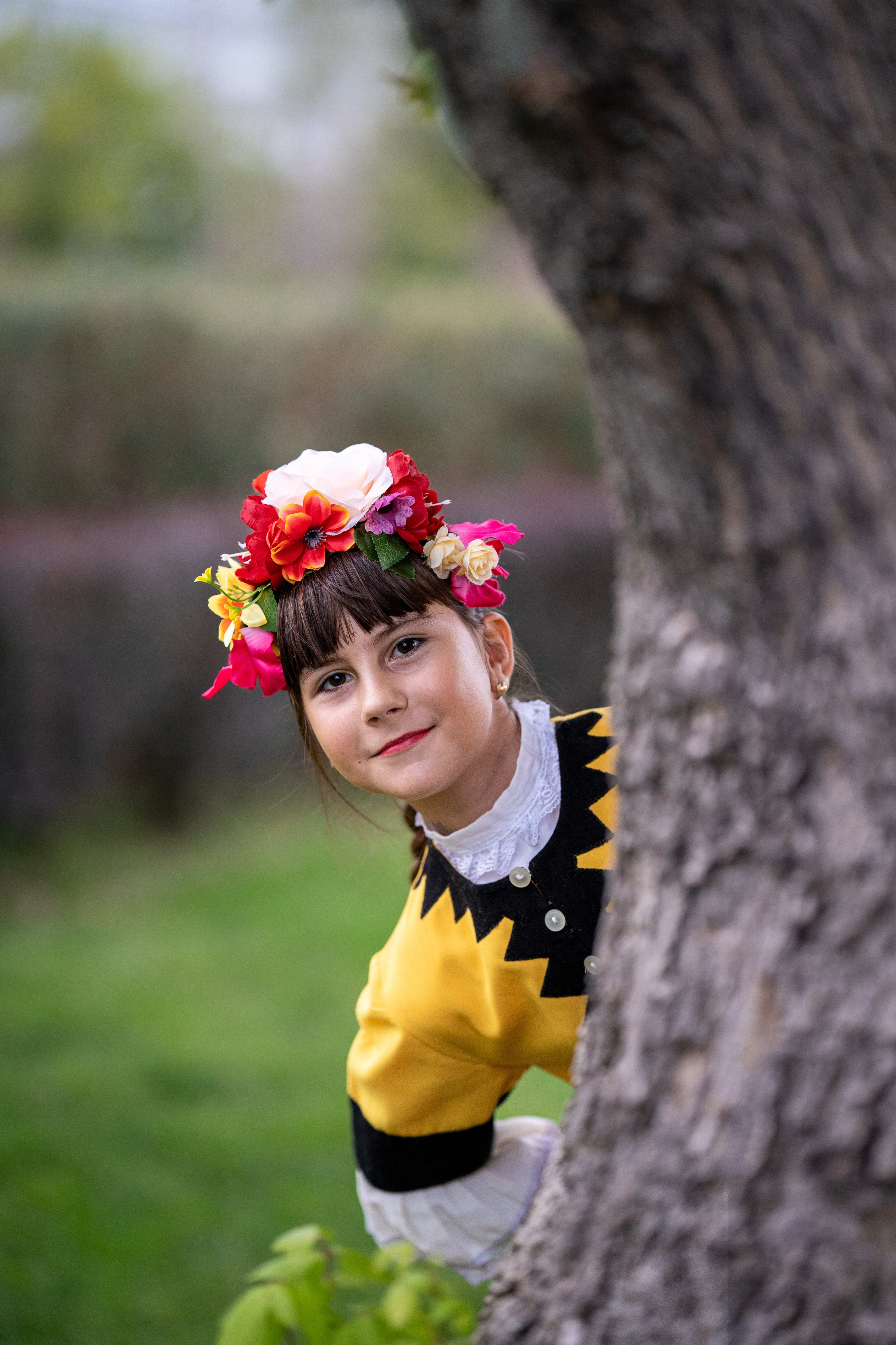 Little girl in traditional folk costume with a flower crown on her head, peeking from behind a tree and smiling.