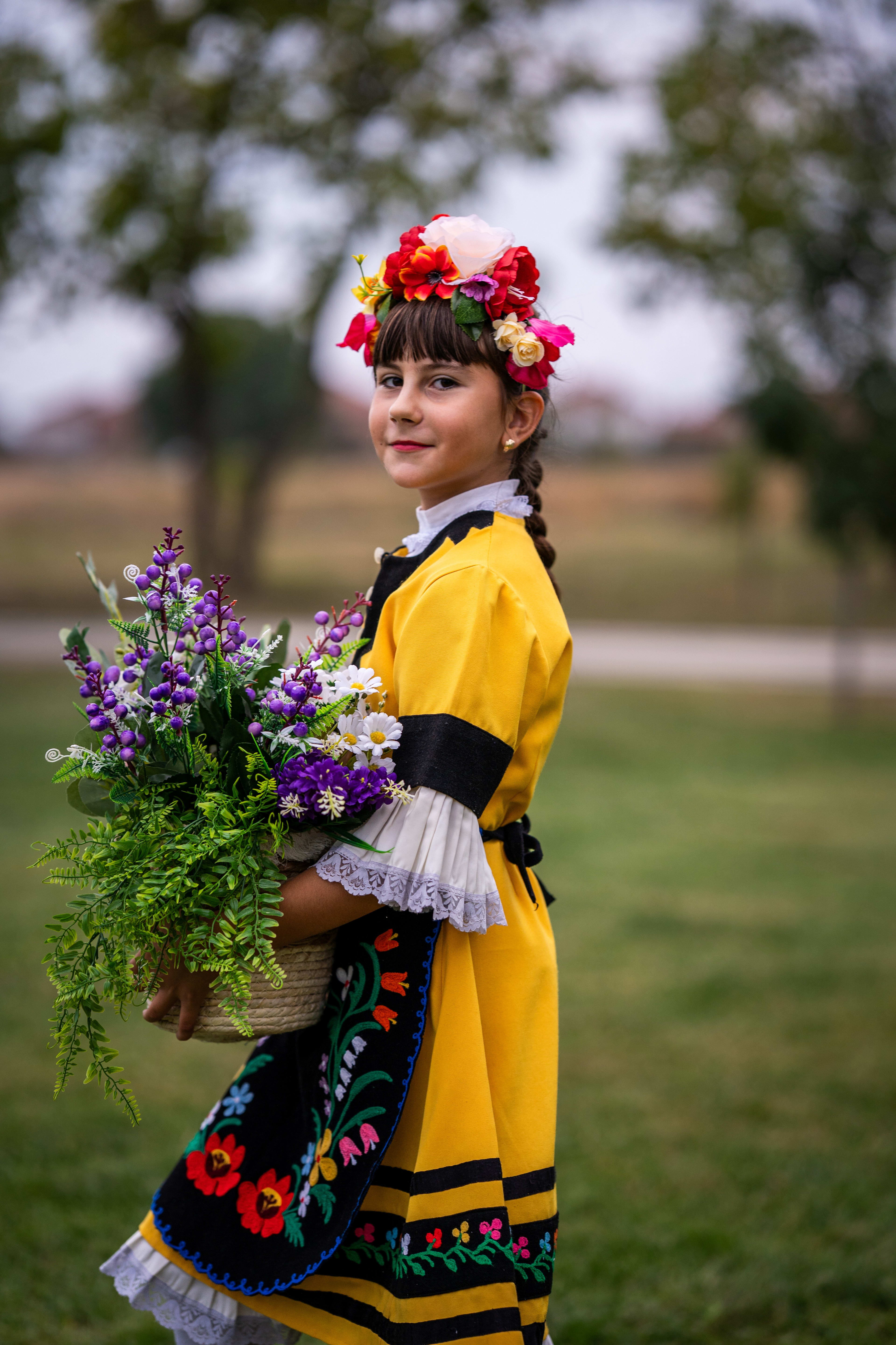 Little girl with a flower crown in traditional folk costume, carrying a basket of flowers and smiling.