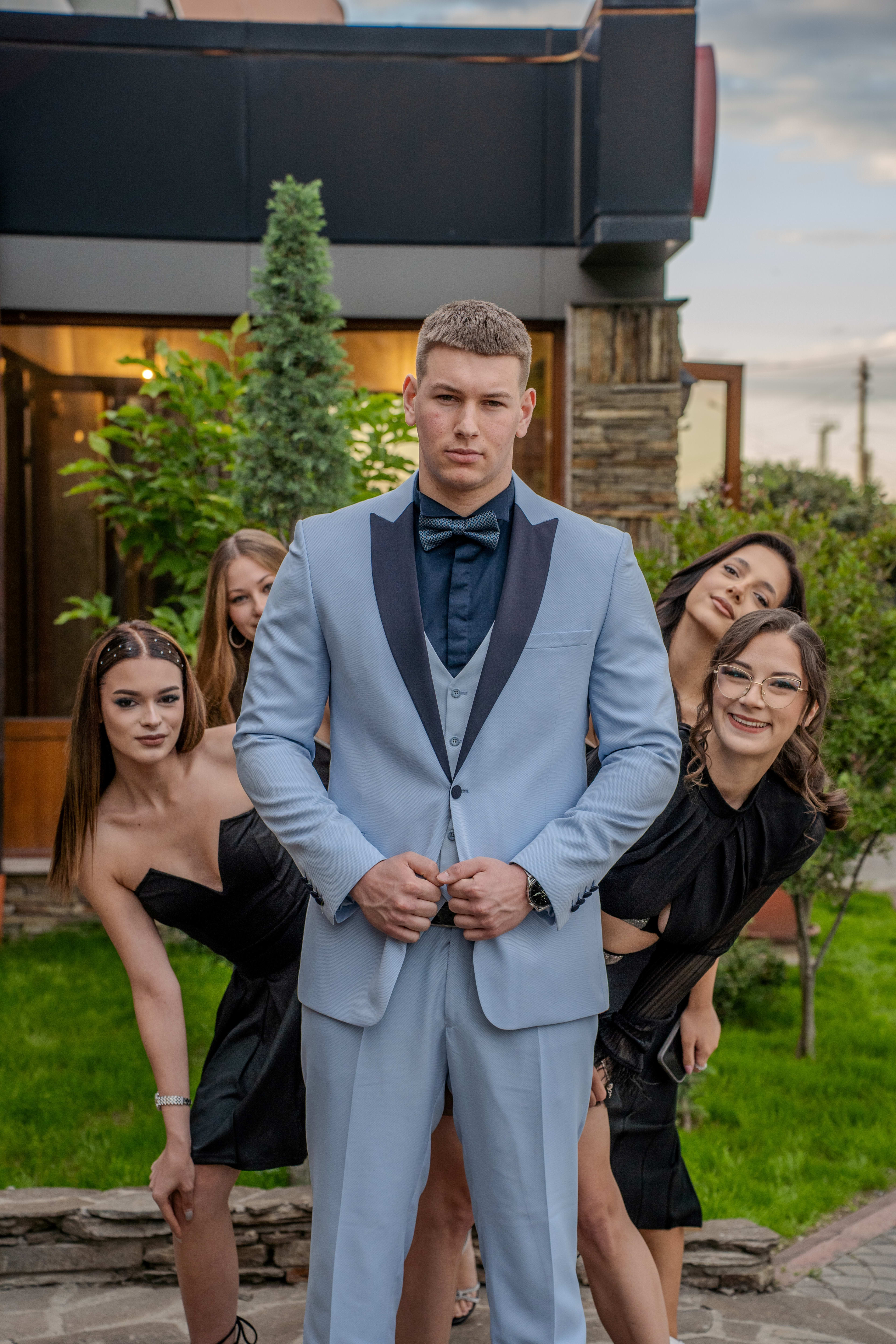A male graduate poses with girls behind him. He is wearing a blue suit, and the girls are in black dresses. Everyone is smiling, with green trees and buildings visible in the background. The girls are peeking out from all sides behind the boy's back.
