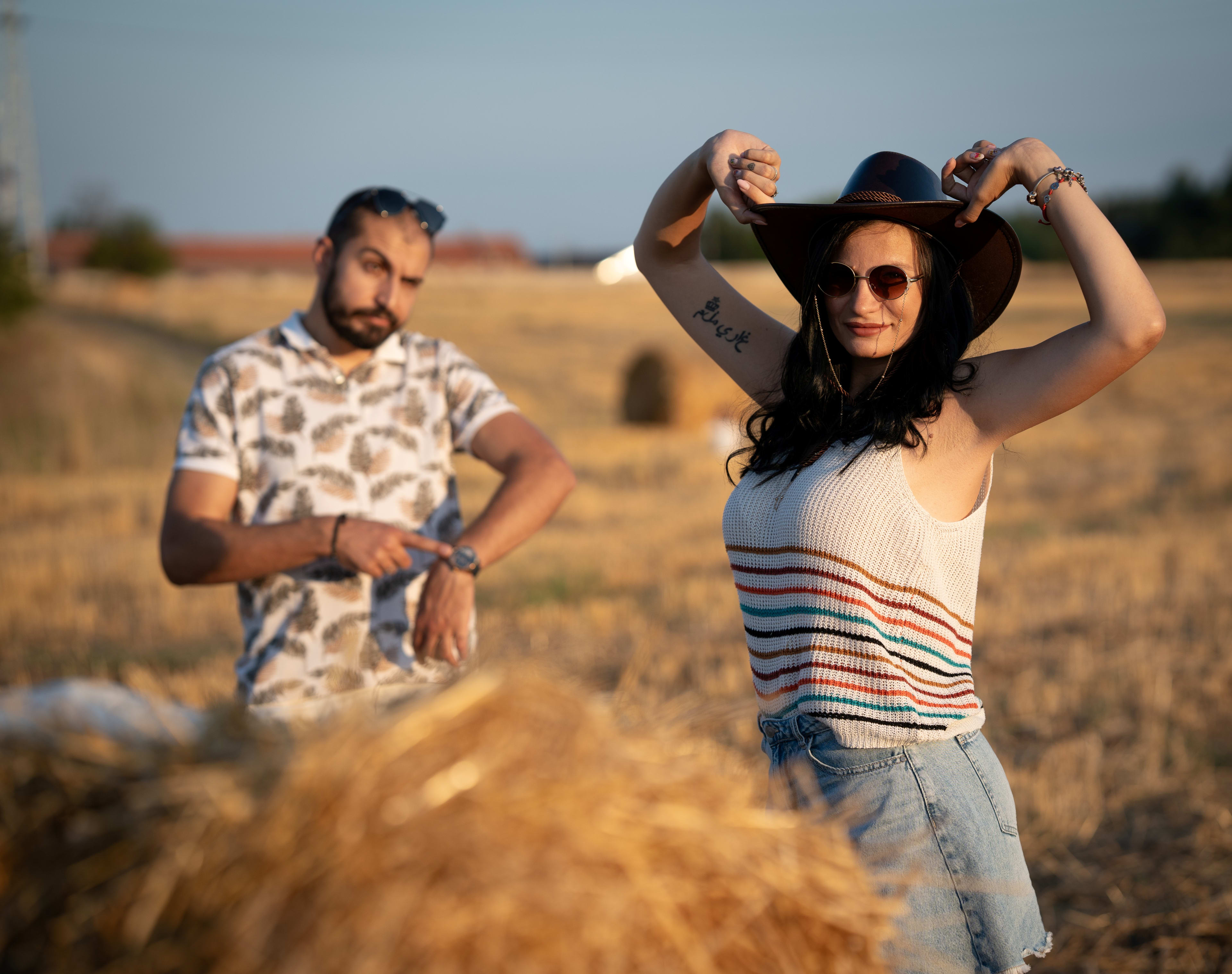 A man points at his watch and looks at his smiling wife. They are dressed in casual clothes and are in a field of straw and straw bales.