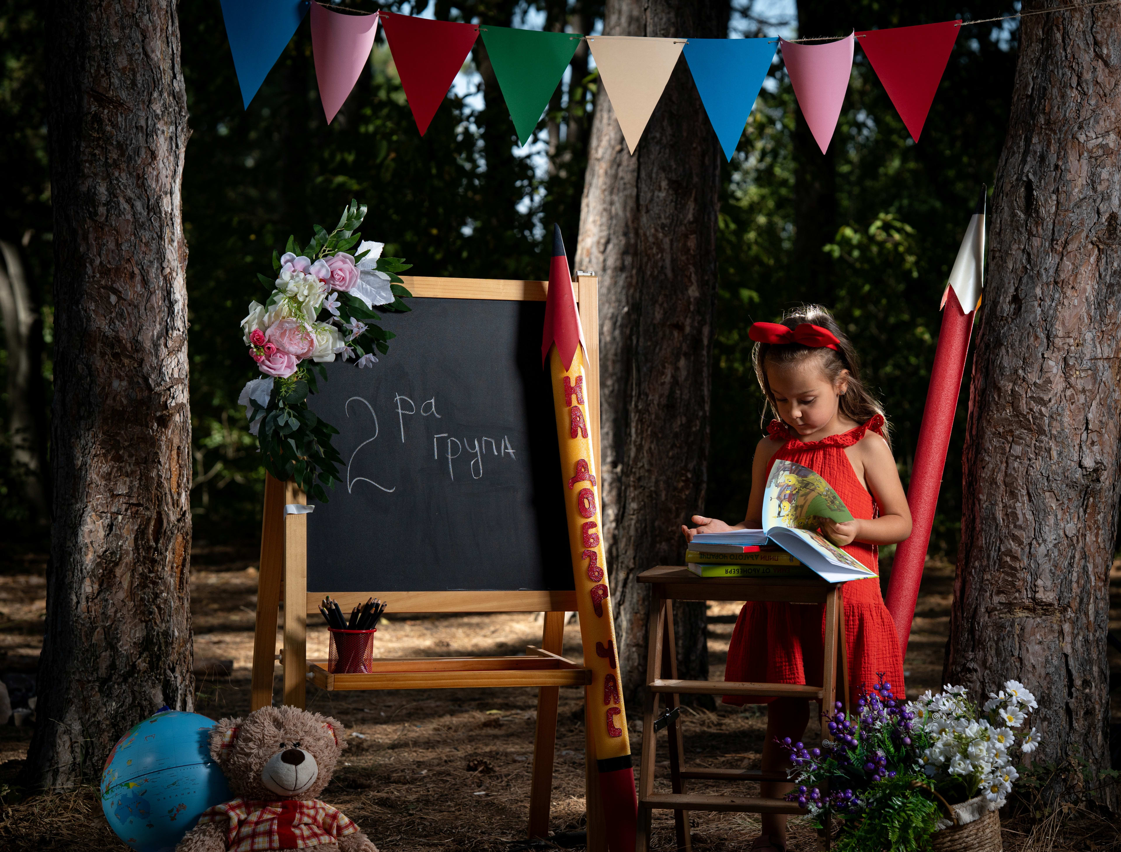 Little girl in a red dress in front of a school set with a chalkboard. On the board it says 'Group 2'. The girl is reading a book.