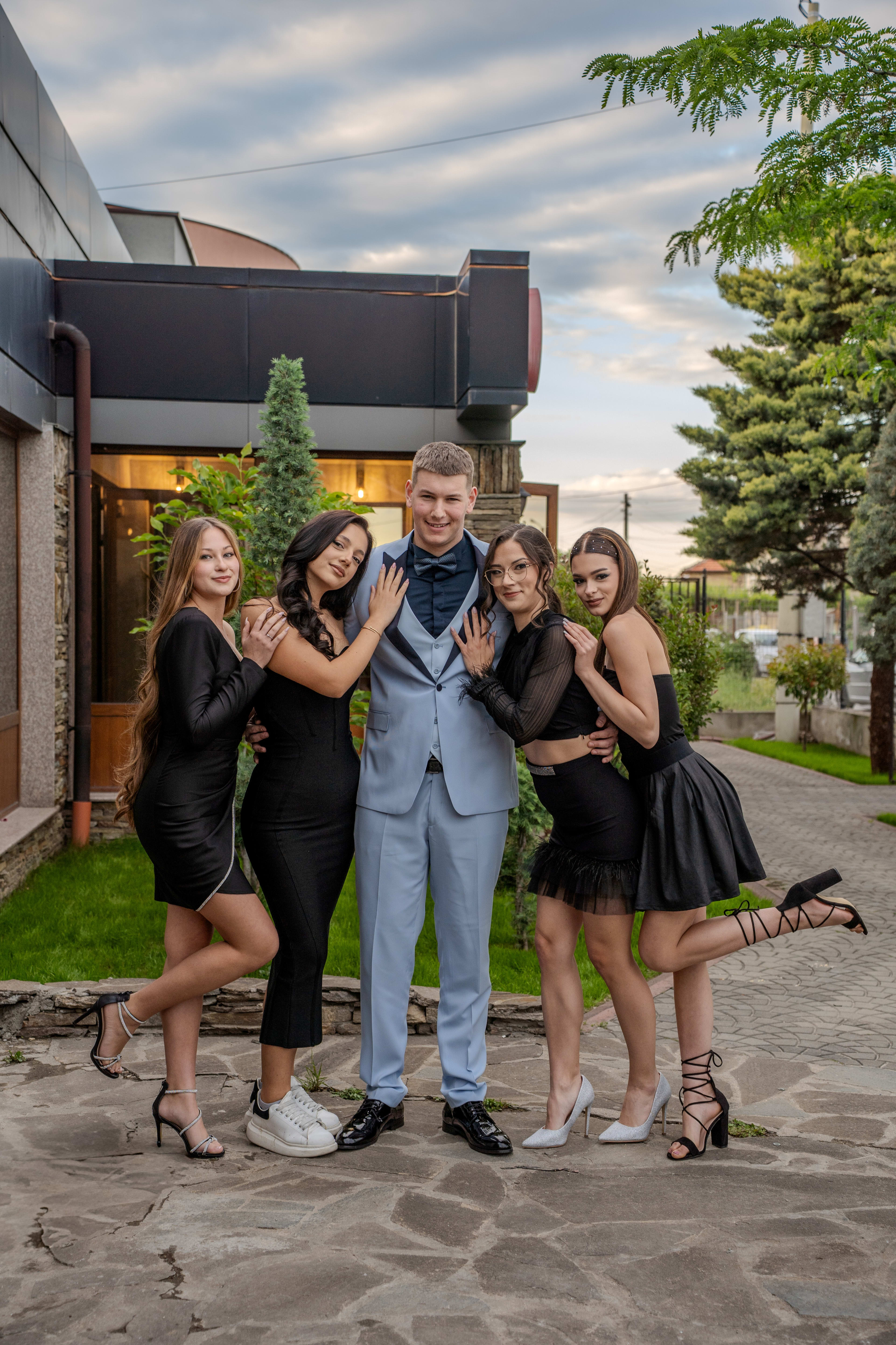 A male graduate poses embraced by four girls, two on each side of him. He is wearing a blue suit, and the girls are in black dresses. Everyone is smiling, with green trees and buildings visible in the background.