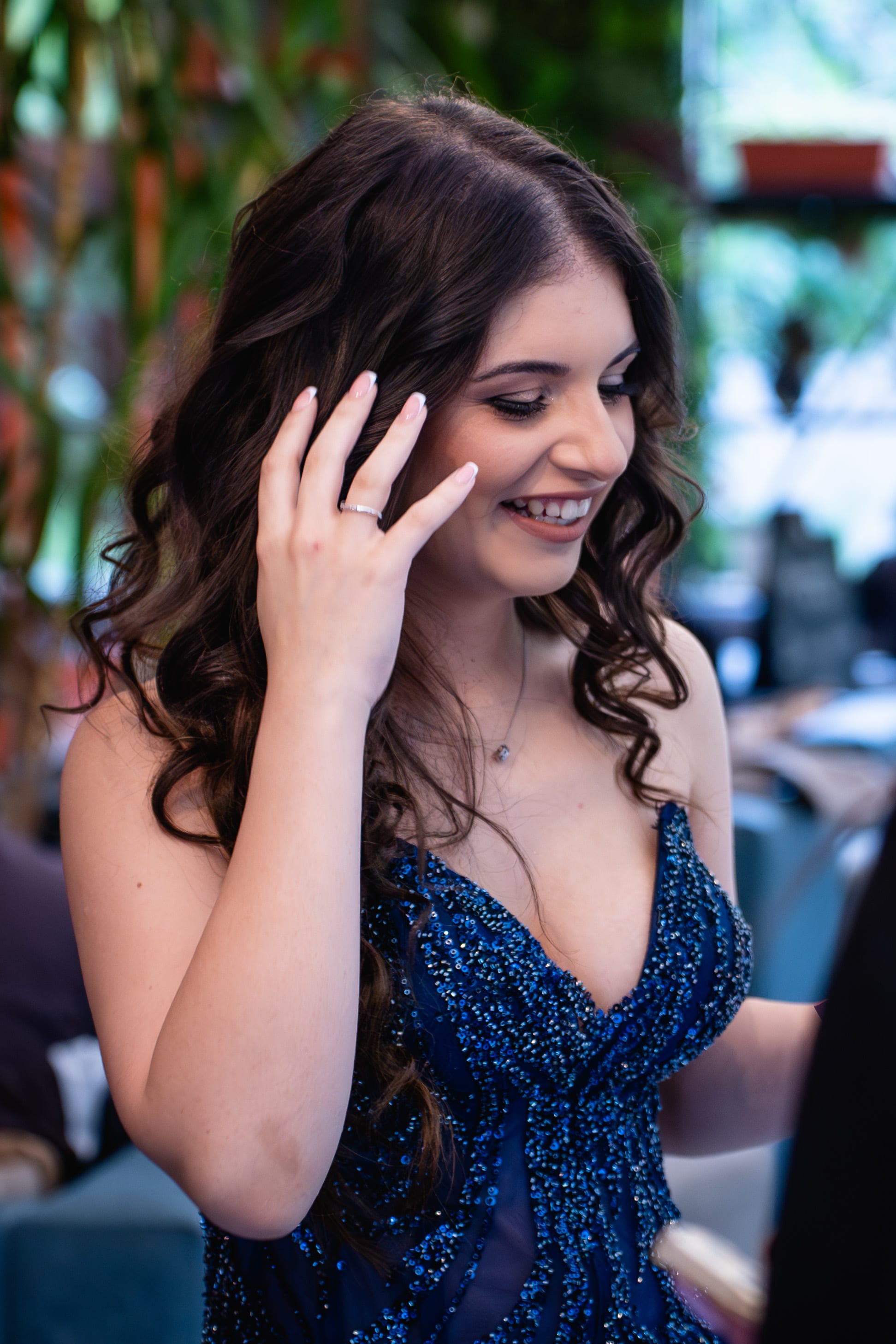 A female graduate in a blue dress is smiling and tucking her hair behind her ear. She has long, slightly wavy hair and is wearing a blue dress with white details.