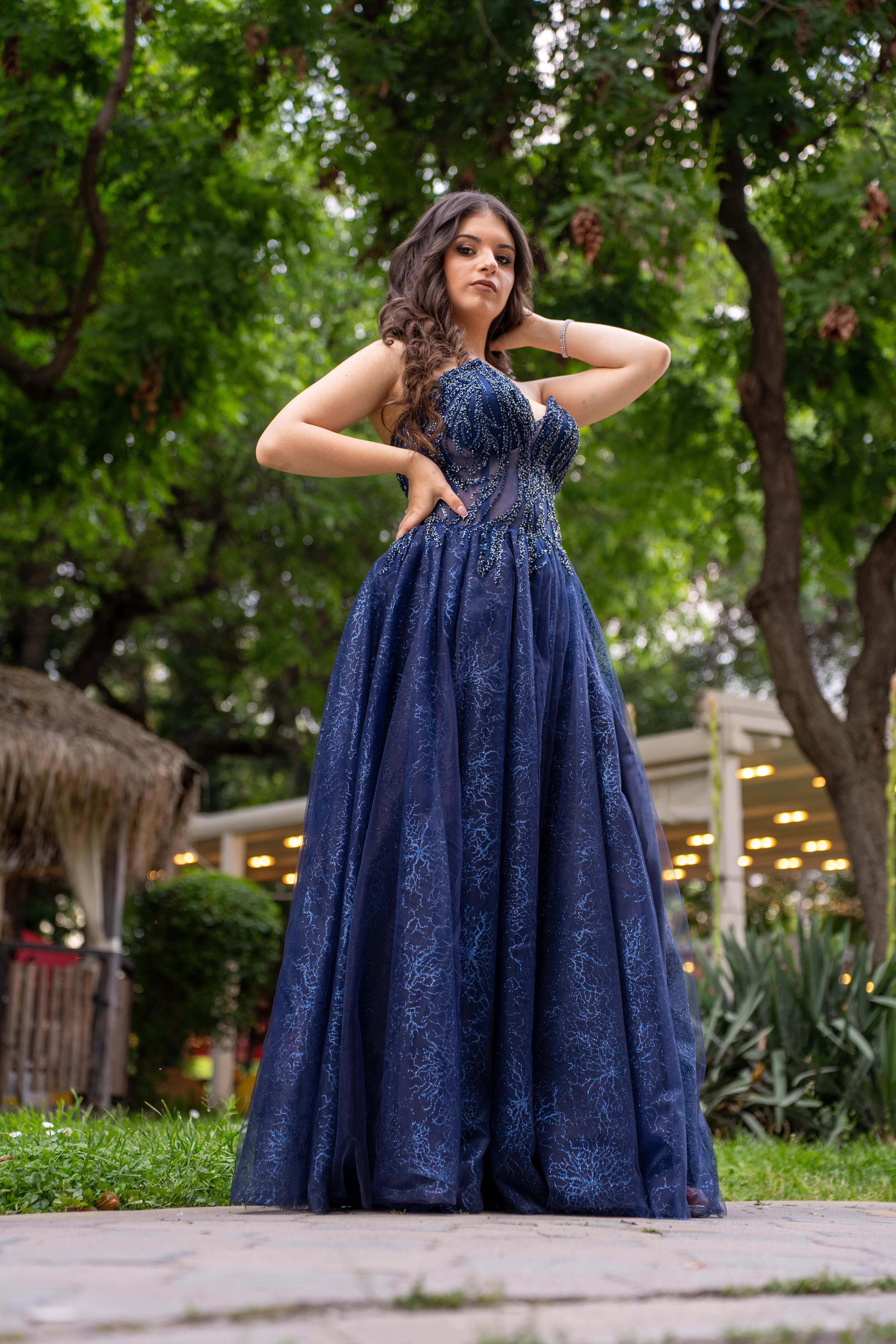 A female graduate in a blue dress, photographed from ground level, as she slightly smiles and looks at the camera. She has long, slightly wavy hair and is wearing a blue dress with white details. Green trees are visible in the background.