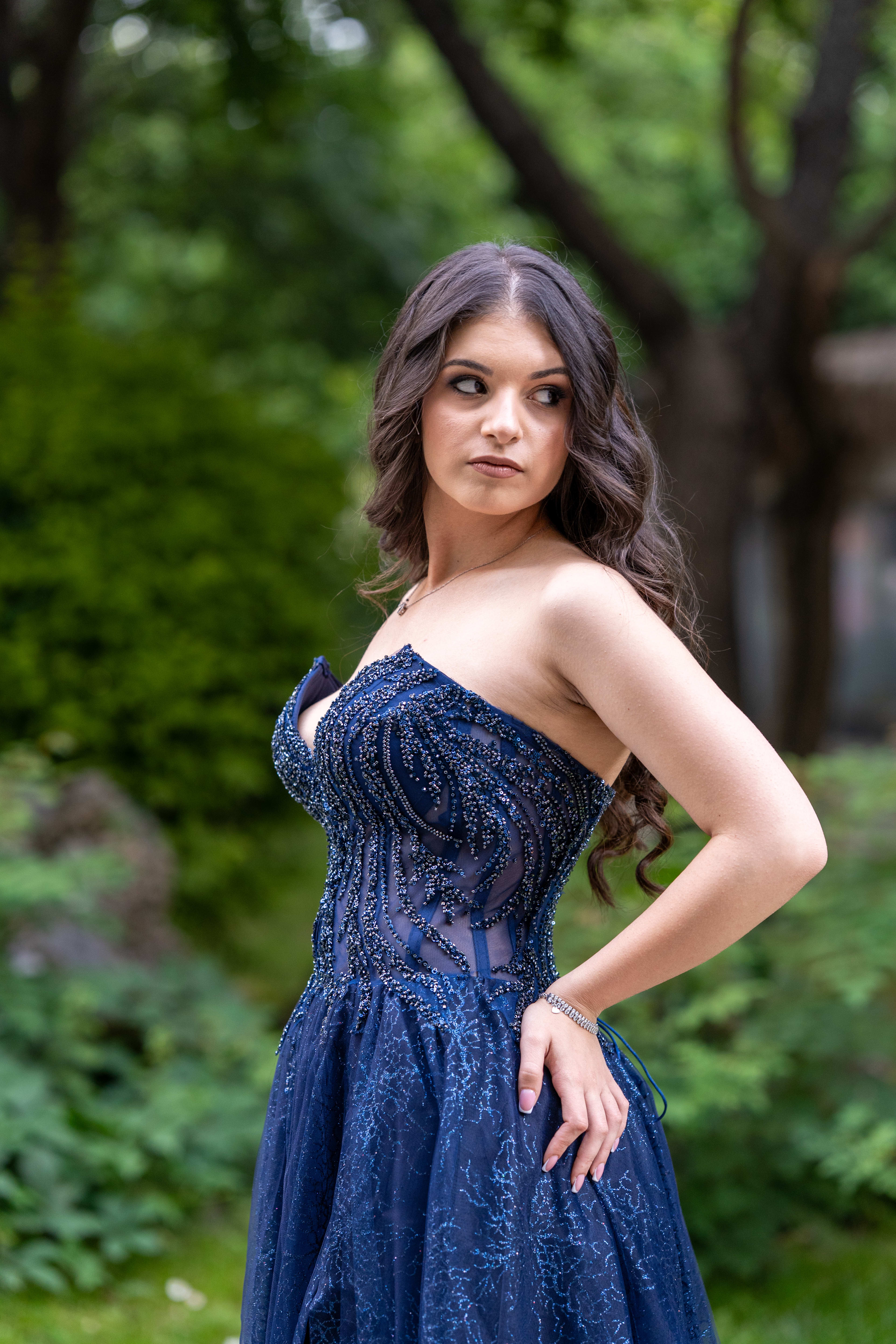 A female graduate in a blue dress poses looking sideways. She has long, slightly wavy hair and is wearing a blue dress with white details. Green trees are visible in the background. Her face is serious, and her gaze is directed towards something in the distance.