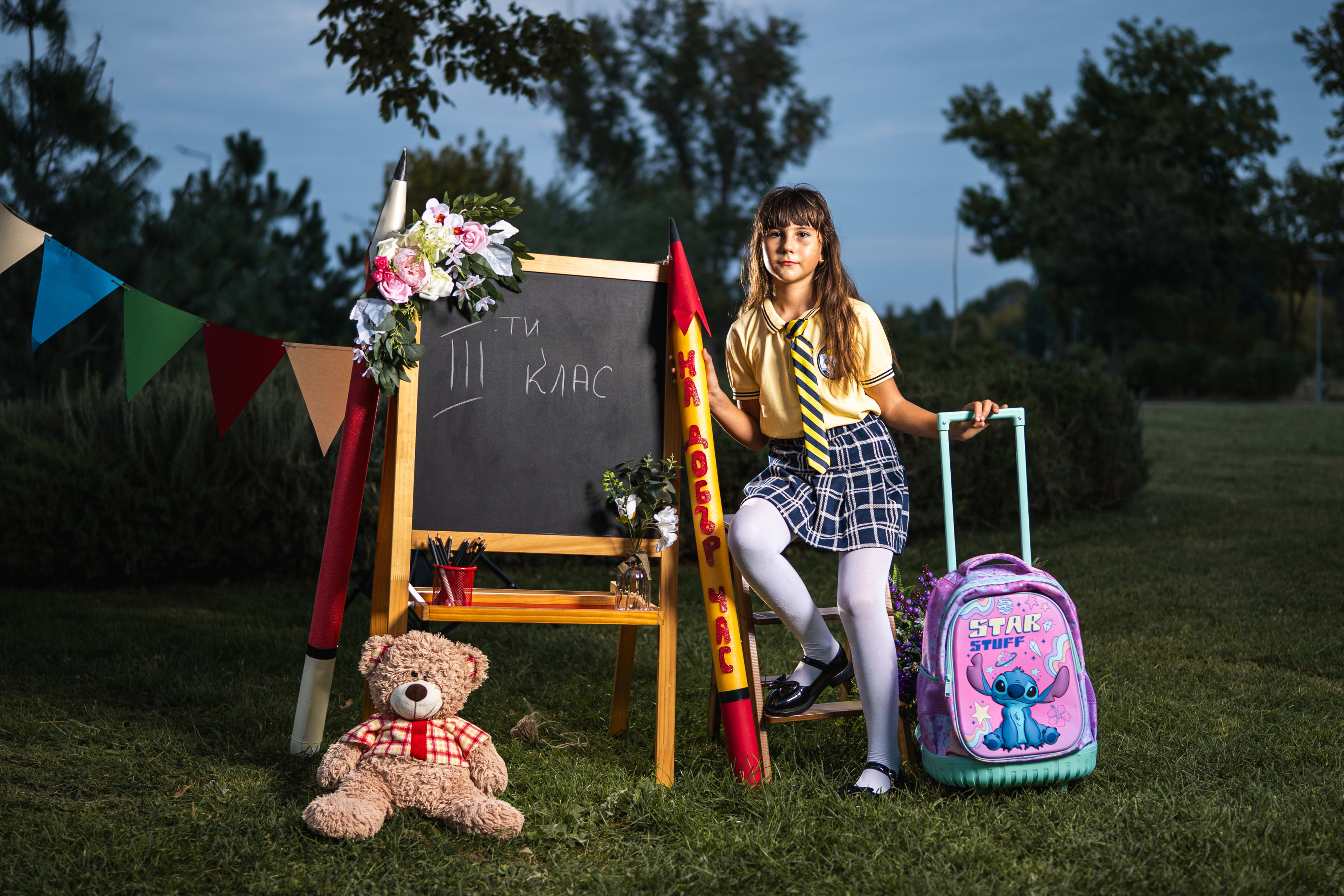Schoolgirl in uniform posing in front of a school set with a chalkboard, a big pencil, a teddy bear, and her backpack. On the board it says '3rd grade'.