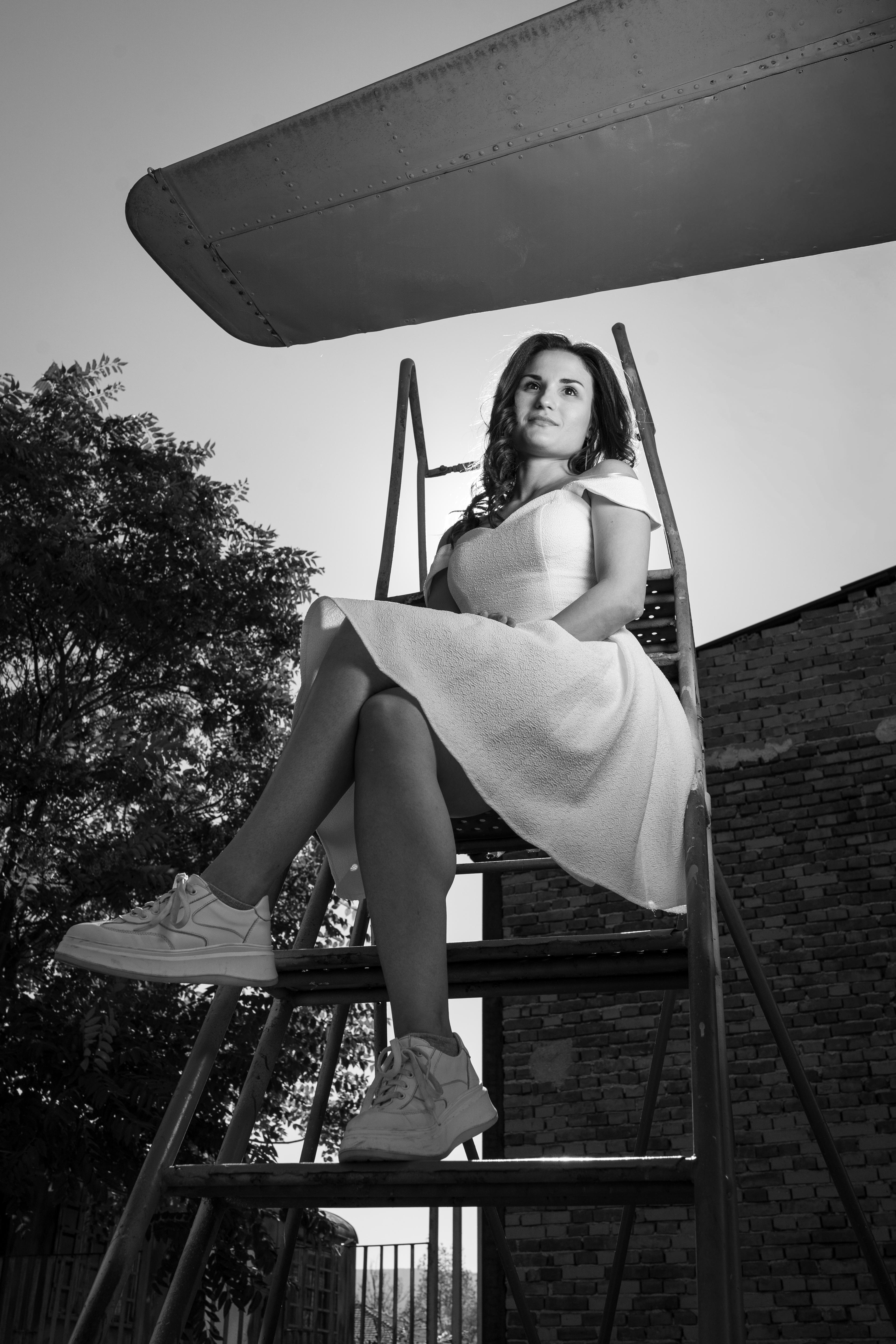 Girl standing on a ladder under the wing of an airplane. She is wearing a white dress and looking at the camera. The photo is black and white.