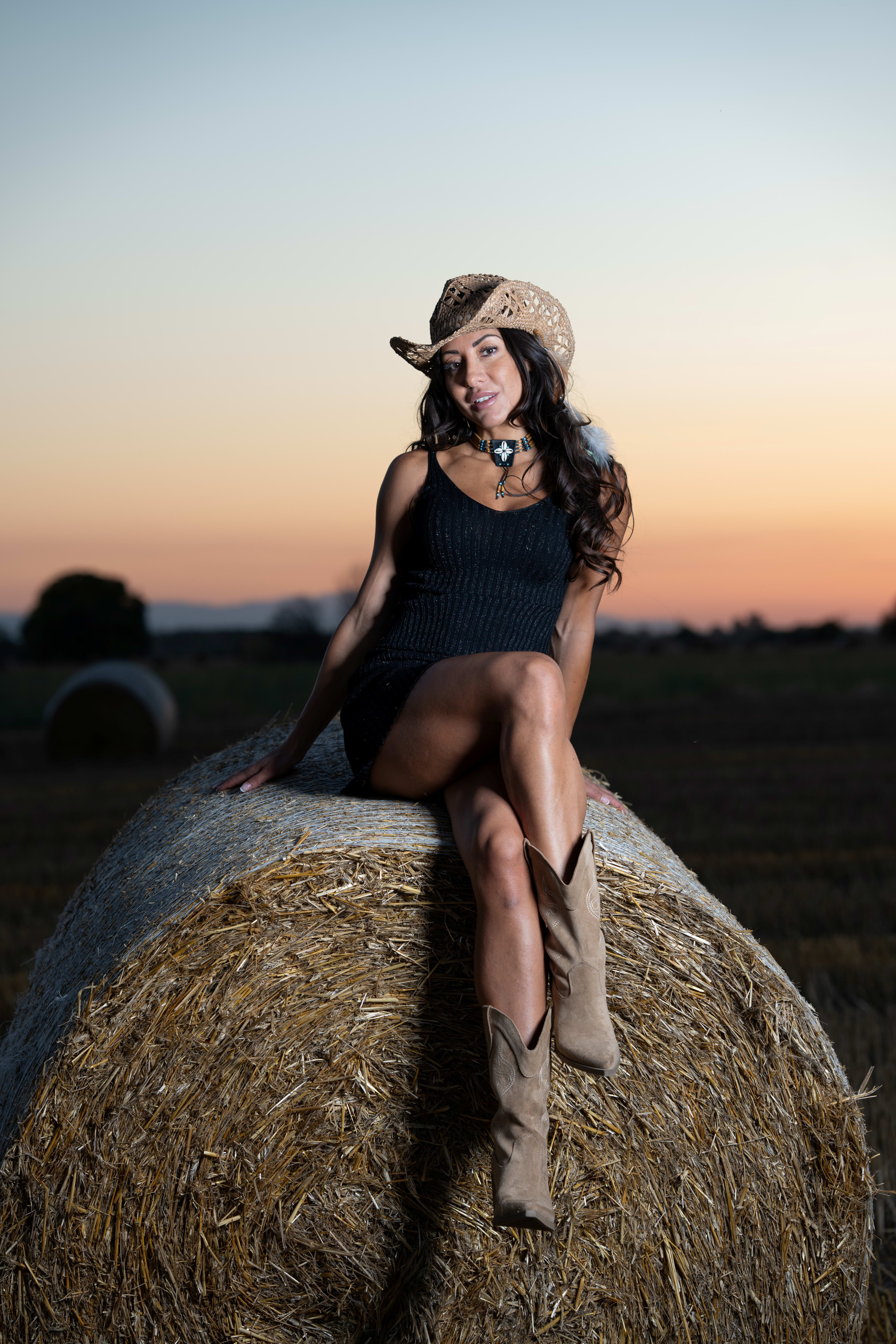 A woman in a black dress with a cowboy hat sits on a straw bale