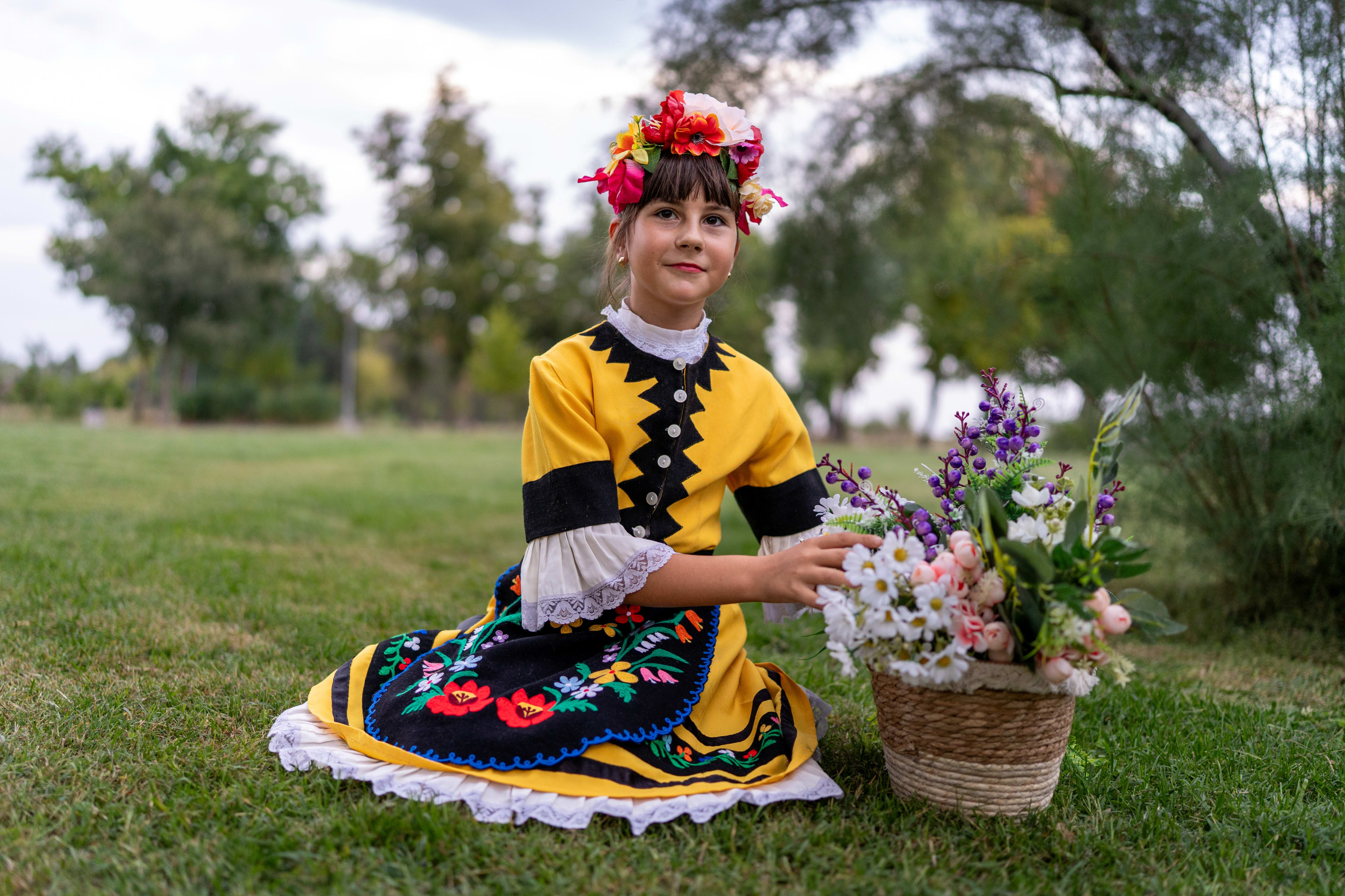 Girl in a traditional folk costume sitting and holding a basket of flowers while smiling.