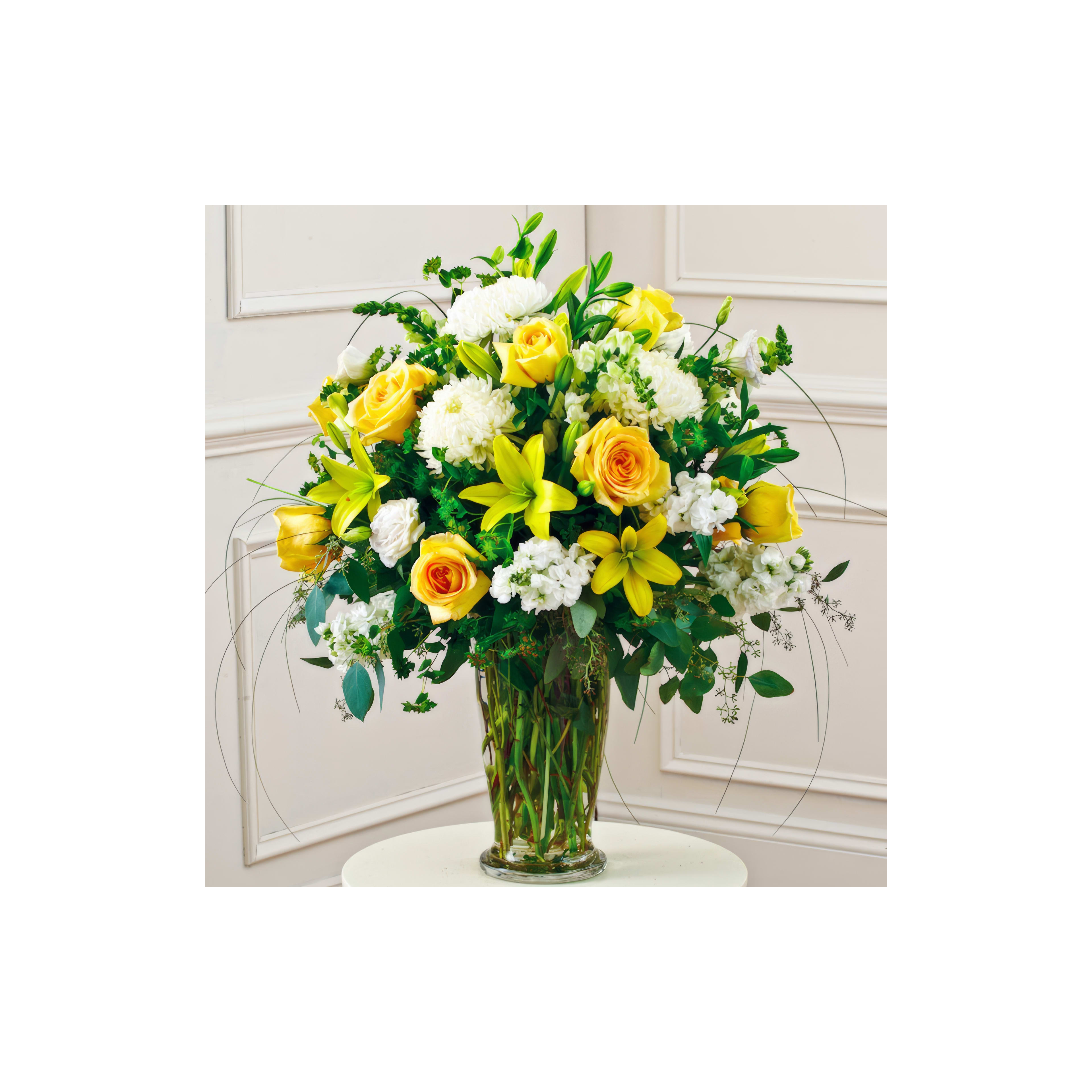 A vibrant arrangement of yellow roses, white lilies, and greenery fills a clear vase on a table.