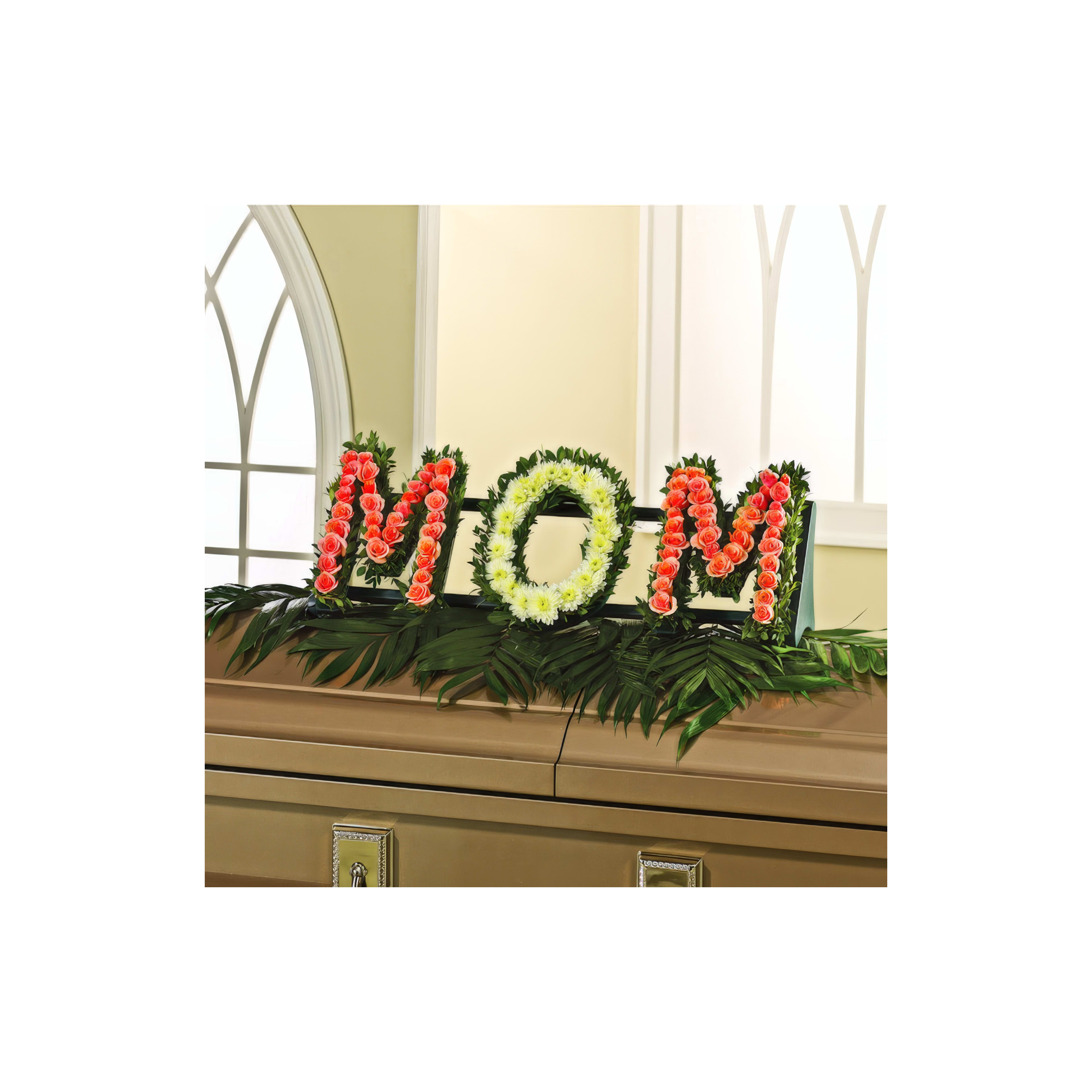 Colorful flowers arranged to spell MOM on a table surrounded by greenery in a well-lit space.
