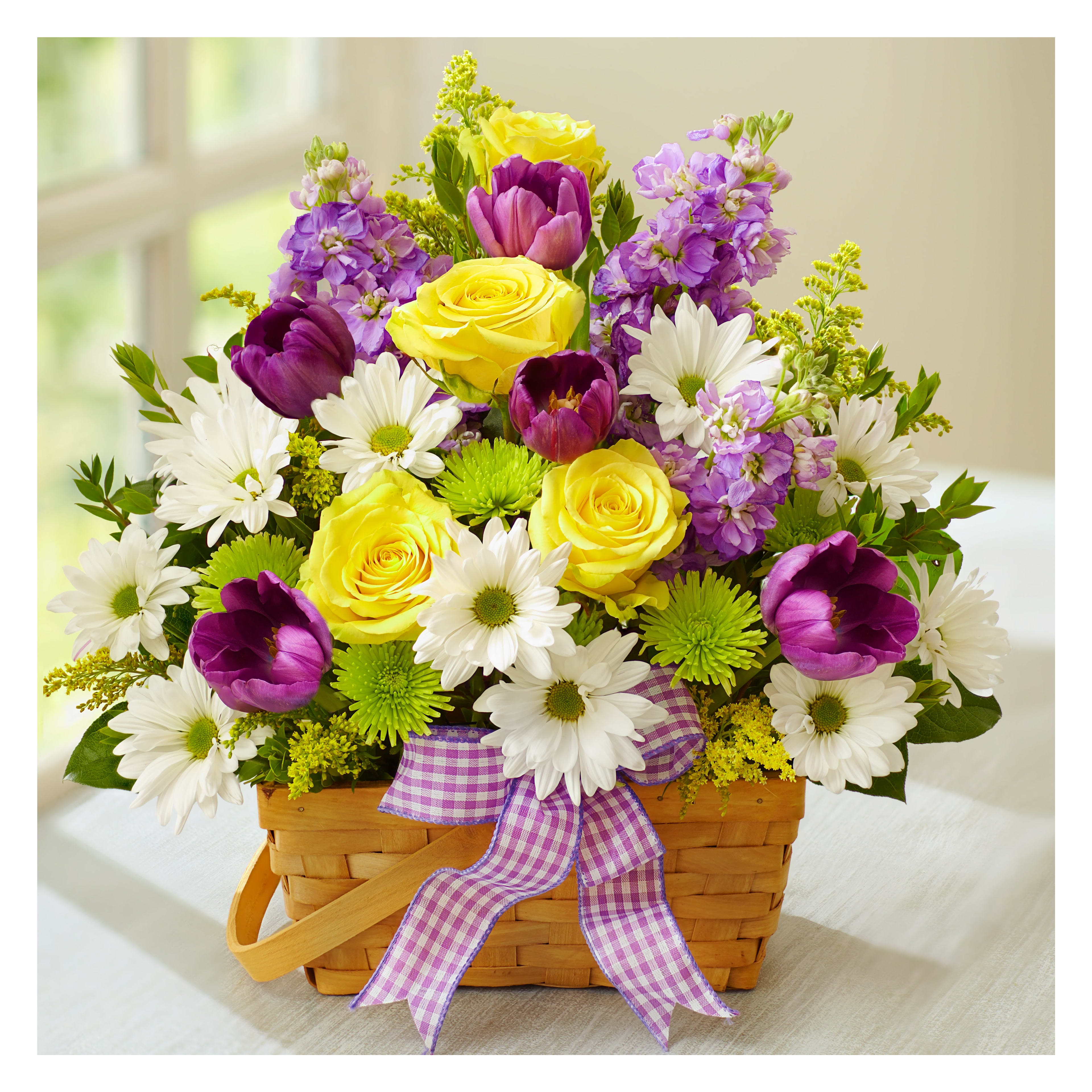 Vibrant basket filled with yellow roses, purple tulips, and white daisies in a bright room.