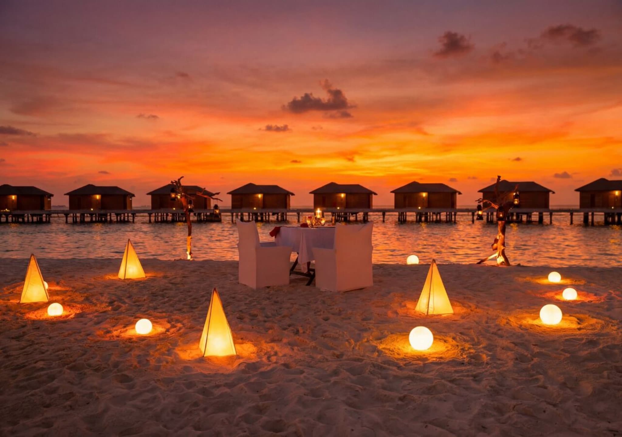Dekket bord på stranda med lykter i sanden og overvannsvillaer i solnedgang på You & Me Maldives