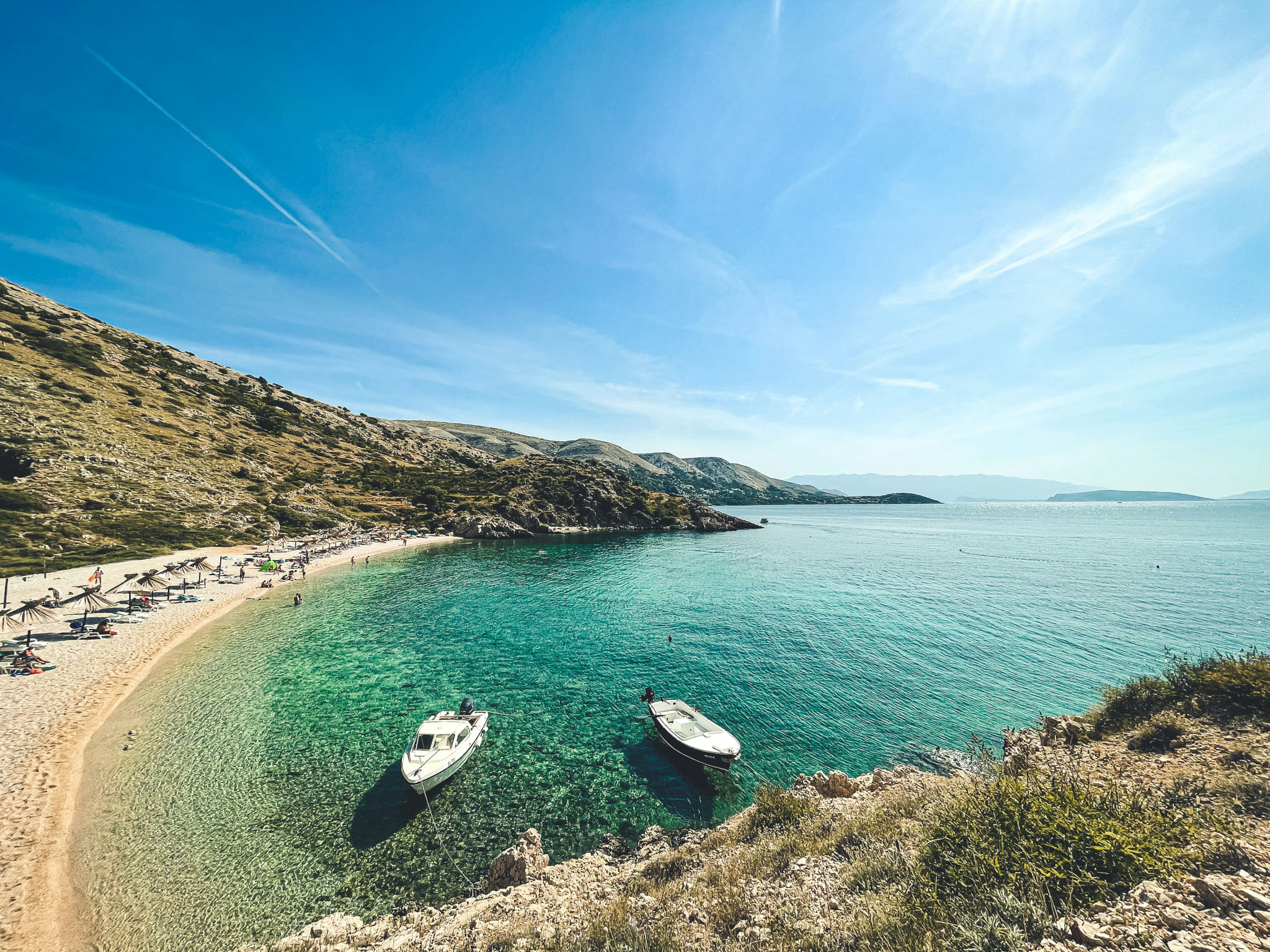 A pale pebble beach in a bay with two boats and clear green water in Croatia