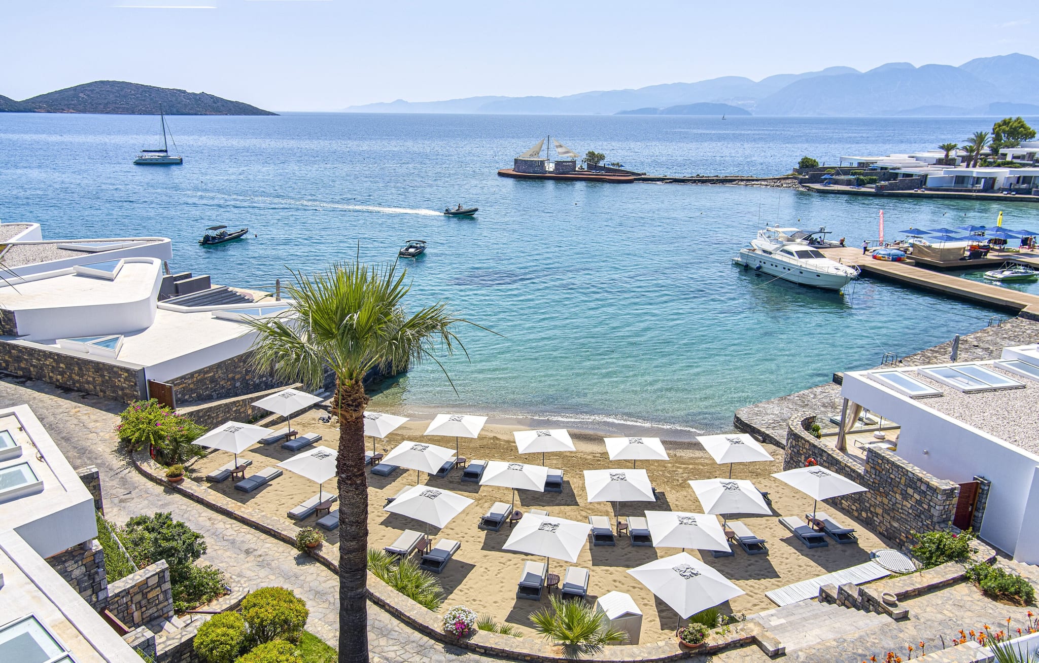 Bukten ved Elounda Beach på Kreta, med parasoller, brygge og fjell over vannet