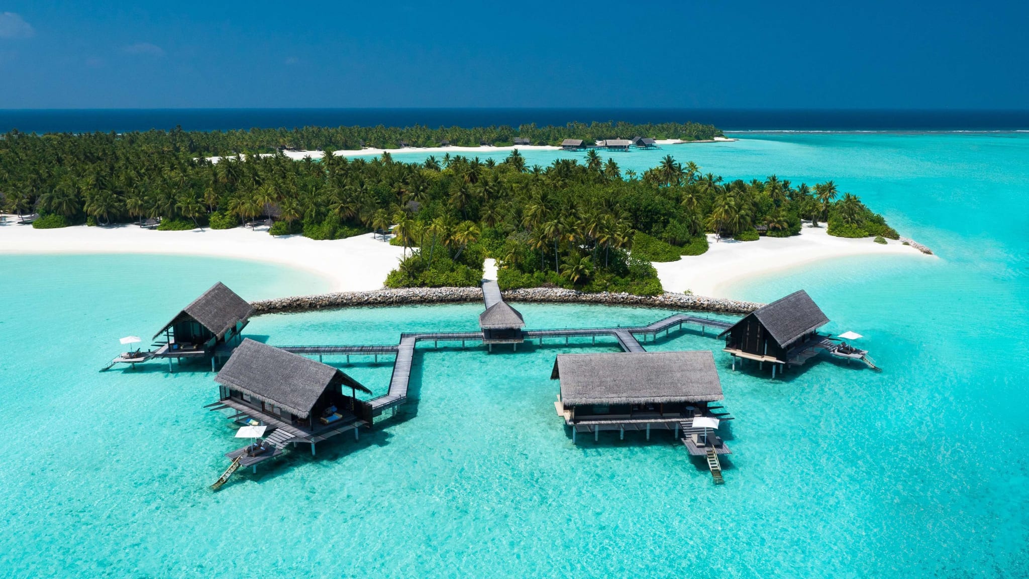 Aerial view of a small Maldives island with white sand, palm trees and overwater villas on stilts in a turquoise lagoon