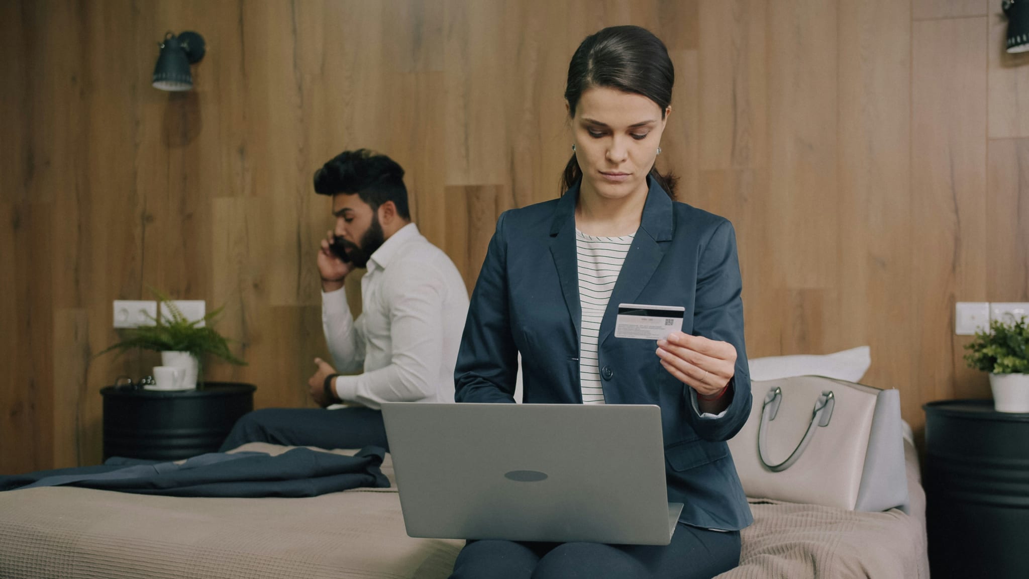 A woman in a hotel room holds a credit card while checking a price on her laptop