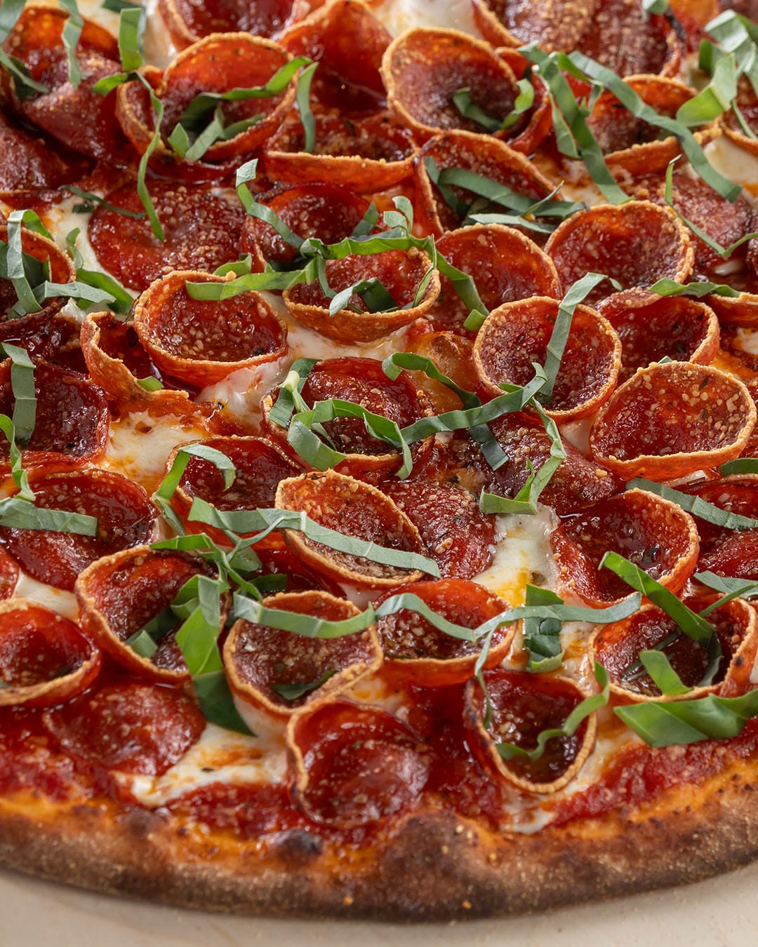 Close up of a round, thin-crust pizza topped with pepperoni slices, melted cheese, tomato sauce, and fresh basil strips. The crust is golden brown and slightly crisp.
