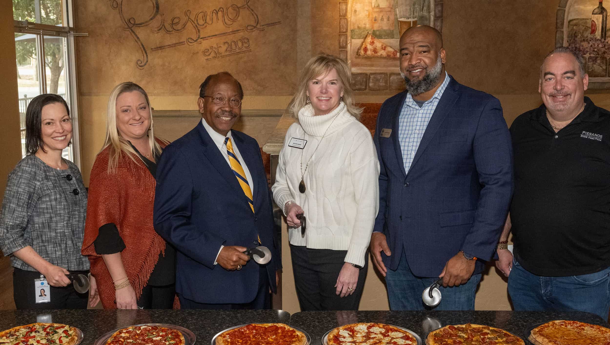 Six smiling people holding pizza cutters, standing in front of a table of 6 different pizzas