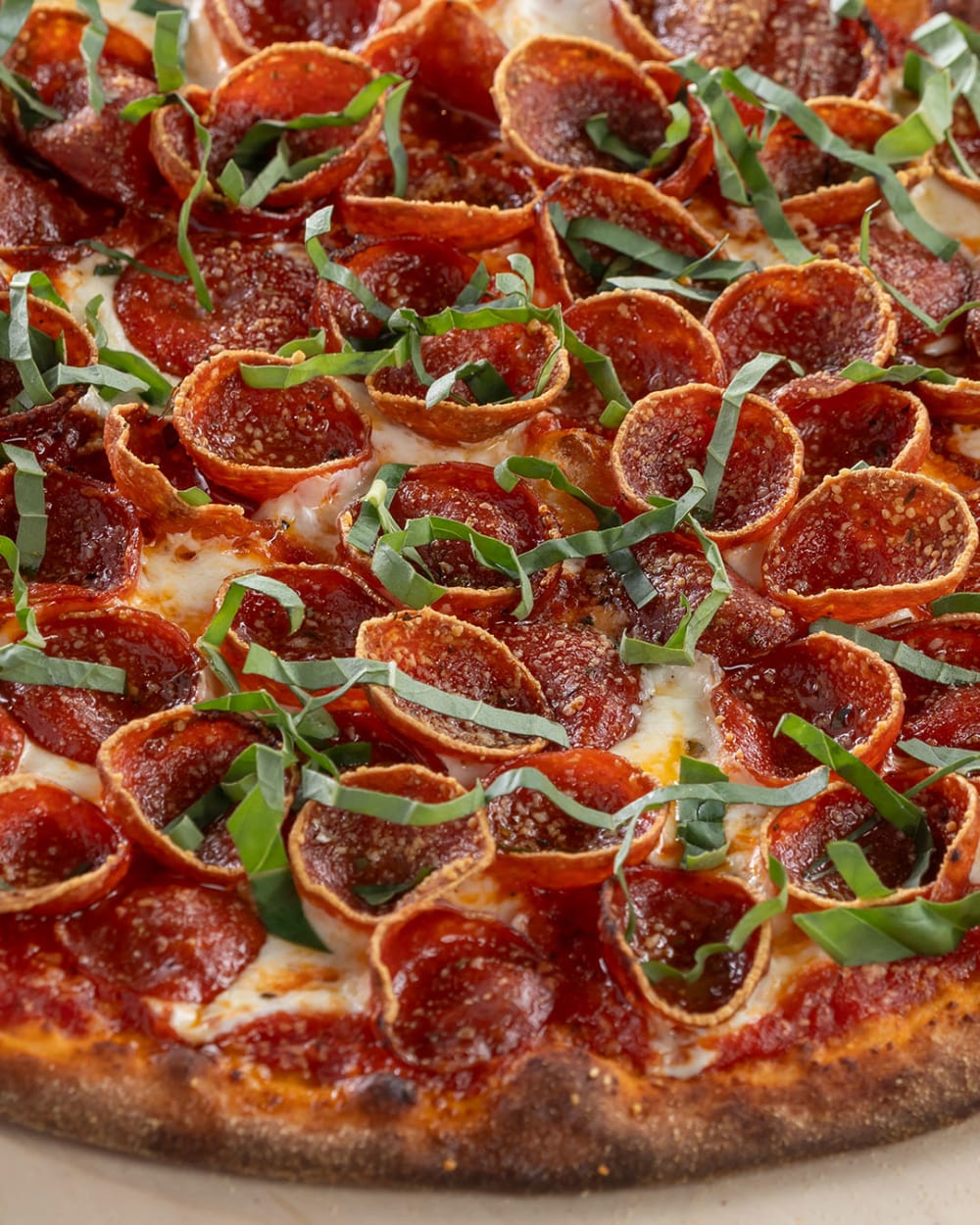 Close up of a round, thin-crust pizza topped with pepperoni slices, melted cheese, tomato sauce, and fresh basil strips. The crust is golden brown and slightly crisp.