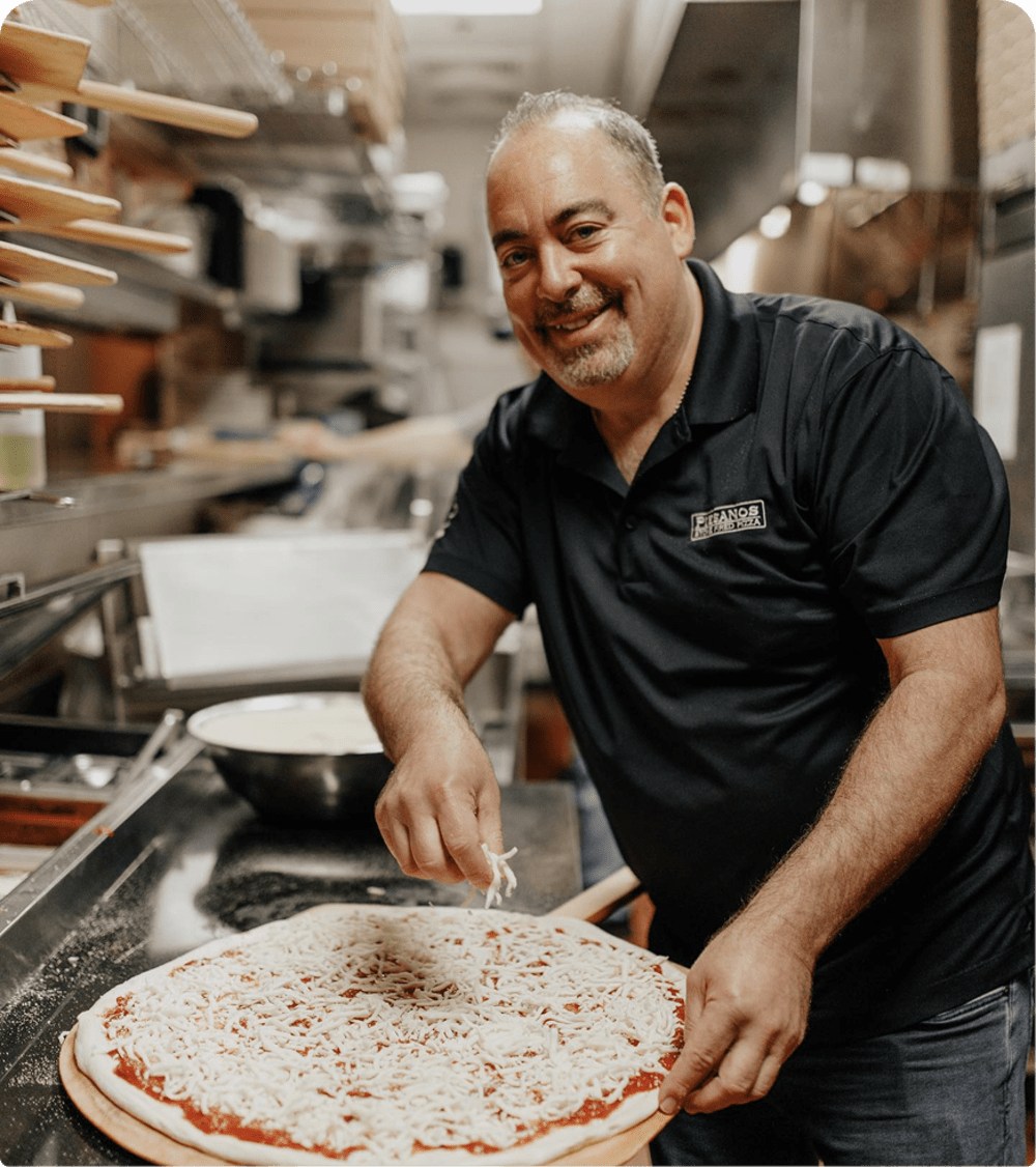 Chef smiling in a black shirt in the kitchen sprinkling cheese on top of pizza