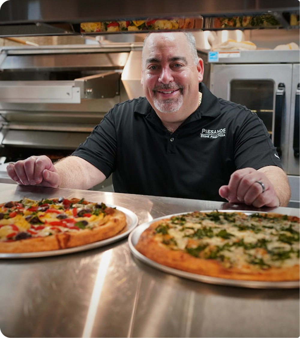 Smiling person standing in the kitchen in front of two different pizzas on silver serving trays