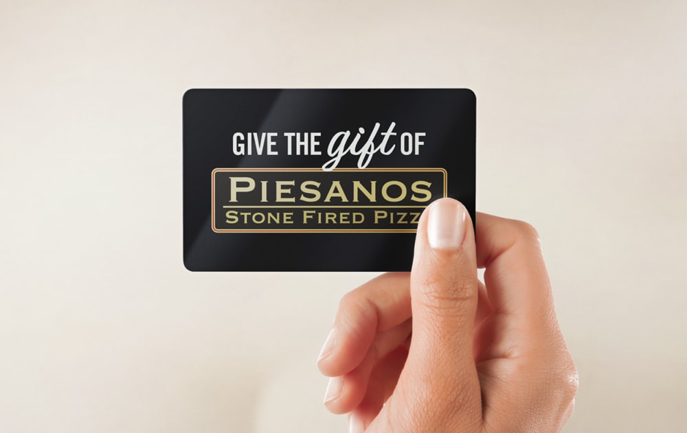 A hand holds a black gift card that reads, Give the gift of Piesanos Stone Fired Pizza in white and gold lettering against a neutral background.