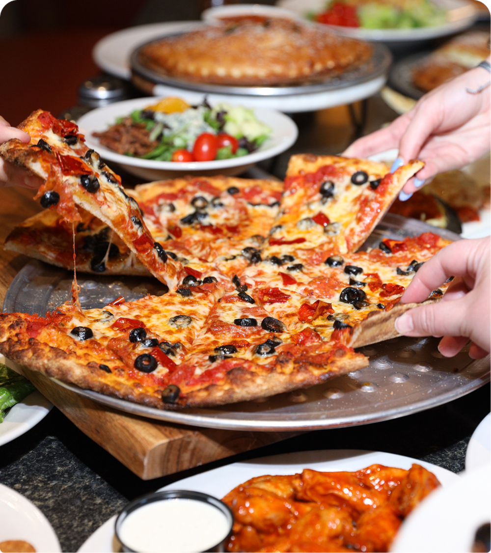 Several hands reach for slices of a large pizza topped with pepperoni and black olives, surrounded by plates of salad, chicken wings with ranch dressing, and a calzone in the background.