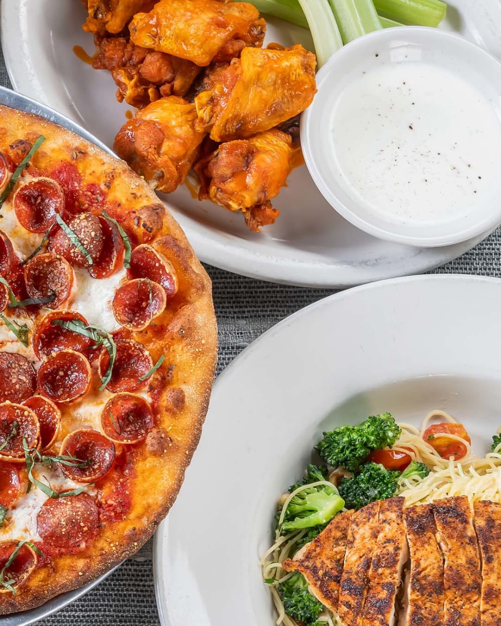 A table with the Antico pizza, buffalo chicken wings with celery and ranch dip, and a plate of grilled chicken breast served over pasta with broccoli and cherry tomatoes.