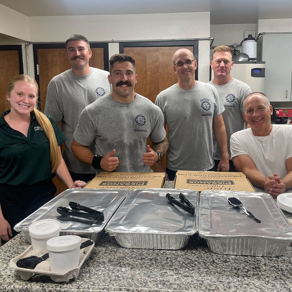 Six smiling people, including five in matching gray shirts and one in a green shirt, stand behind a counter with trays of food, utensils, and takeout containers in a kitchen setting.