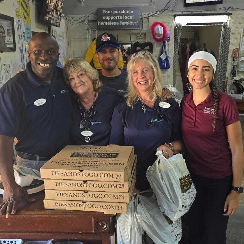 Five smiling people pose together inside a shop, with stacked pizza boxes and Piesanos takeout bags.