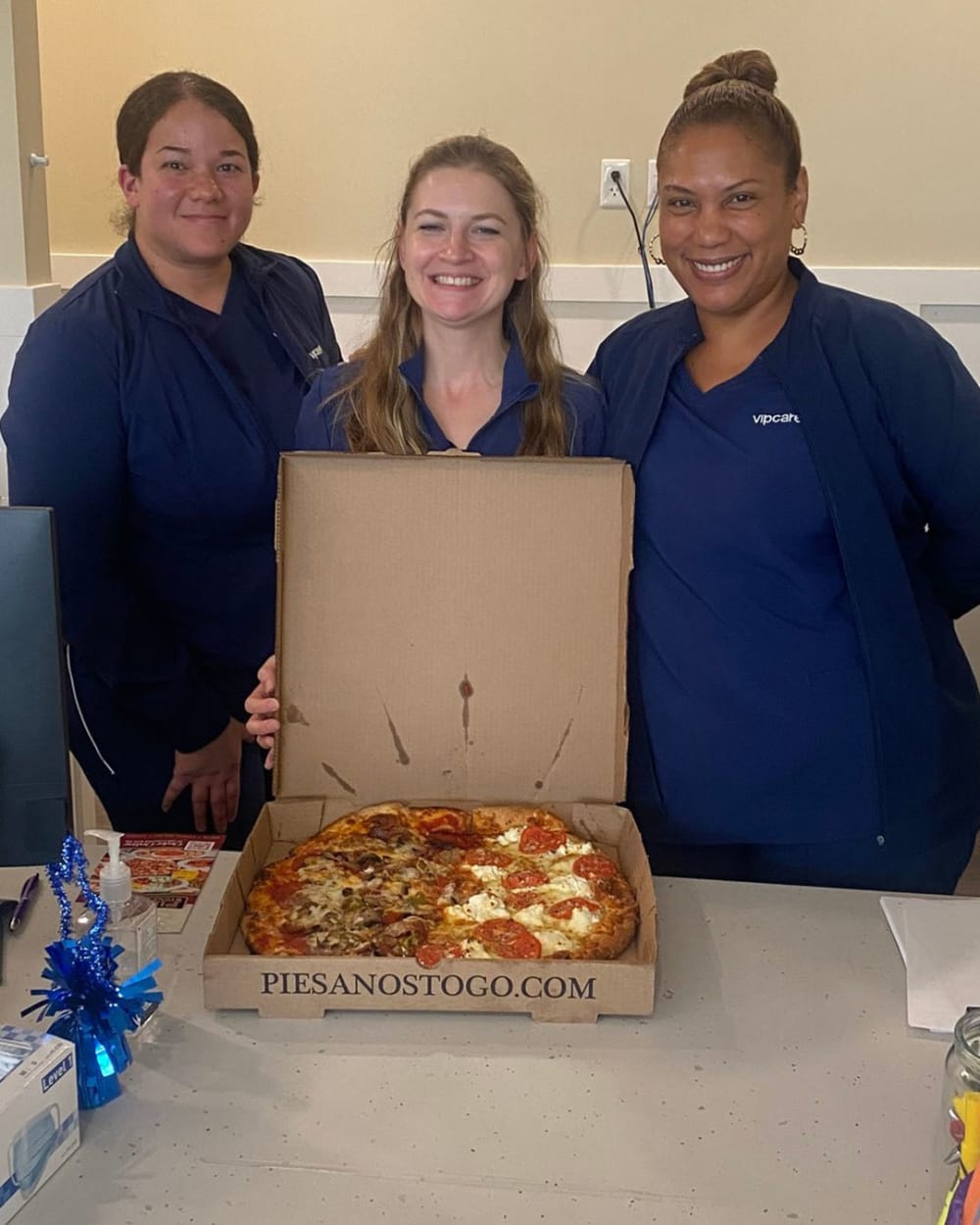 Three women in blue uniforms smile behind a table with an open pizza box displaying a large pizza. The box reads PIESANOSTOGO.COM and there are some snacks and decorations on the table.