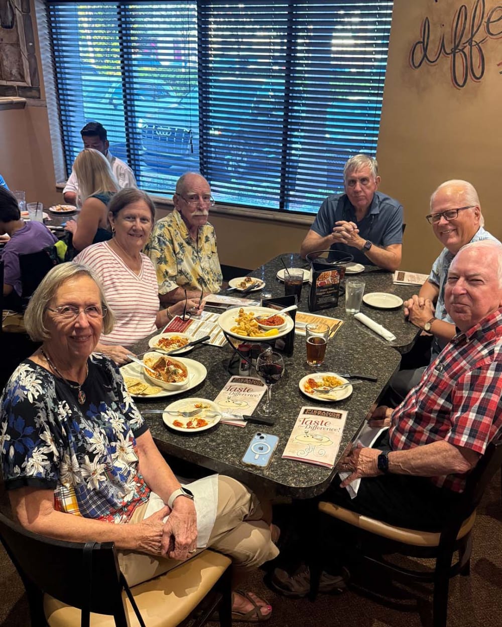Six older adults sit around a table at a restaurant, dining and smiling at the camera. Plates of food, drinks, and menus are on the table, and a window with blinds is in the background.