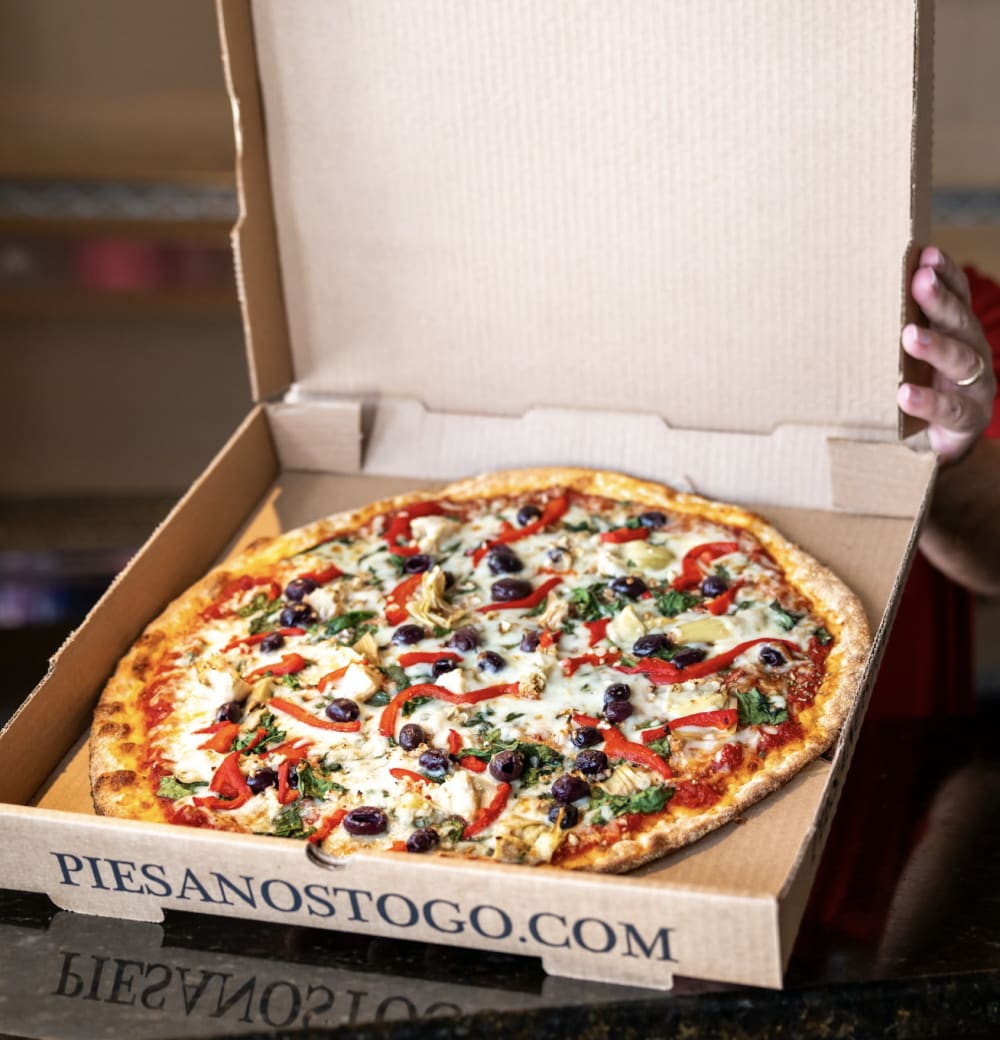 A hand holds open a pizza box revealing a large pizza topped with cheese, red peppers, olives, spinach, and onions. The box has the website PIESANOSTOGO.COM printed on the edge.