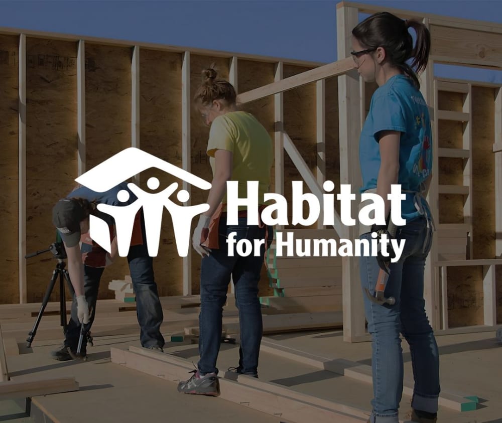 Three people work on building a wooden house frame outdoors. The Habitat for Humanity logo and text are overlaid on the image. Two people hold tools, and one uses a tape measure.