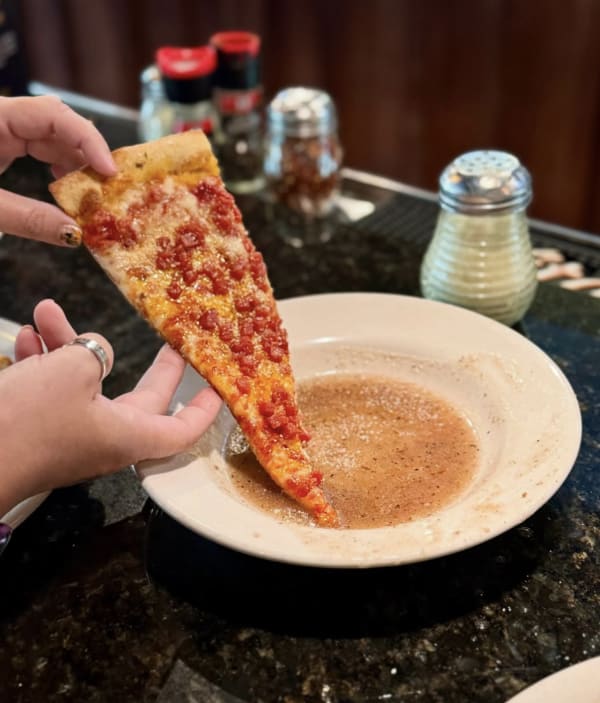 Two hands holding a slice of pizza dipping it into a bowl of sauce on a counter