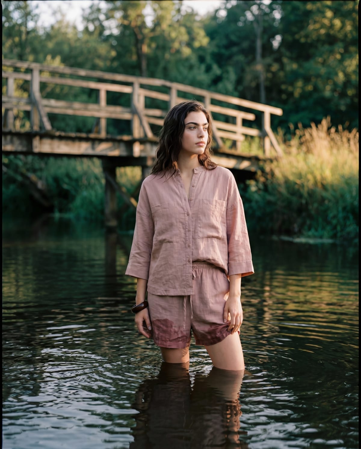 Modelo de camisa e short de linho rosa com água até o joelho num rio sob uma ponte de madeira