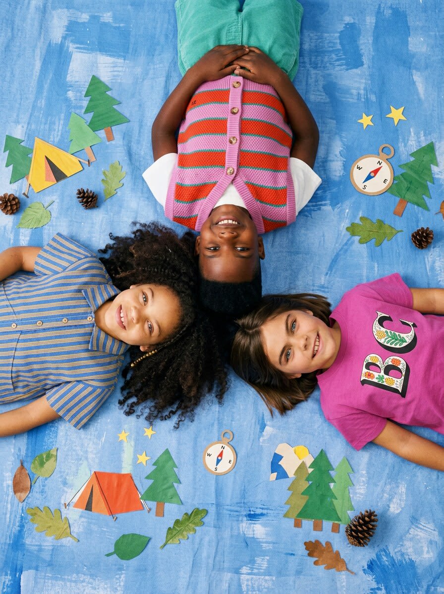 Three children in striped and pink kidswear lying head to head among paper tents and pines