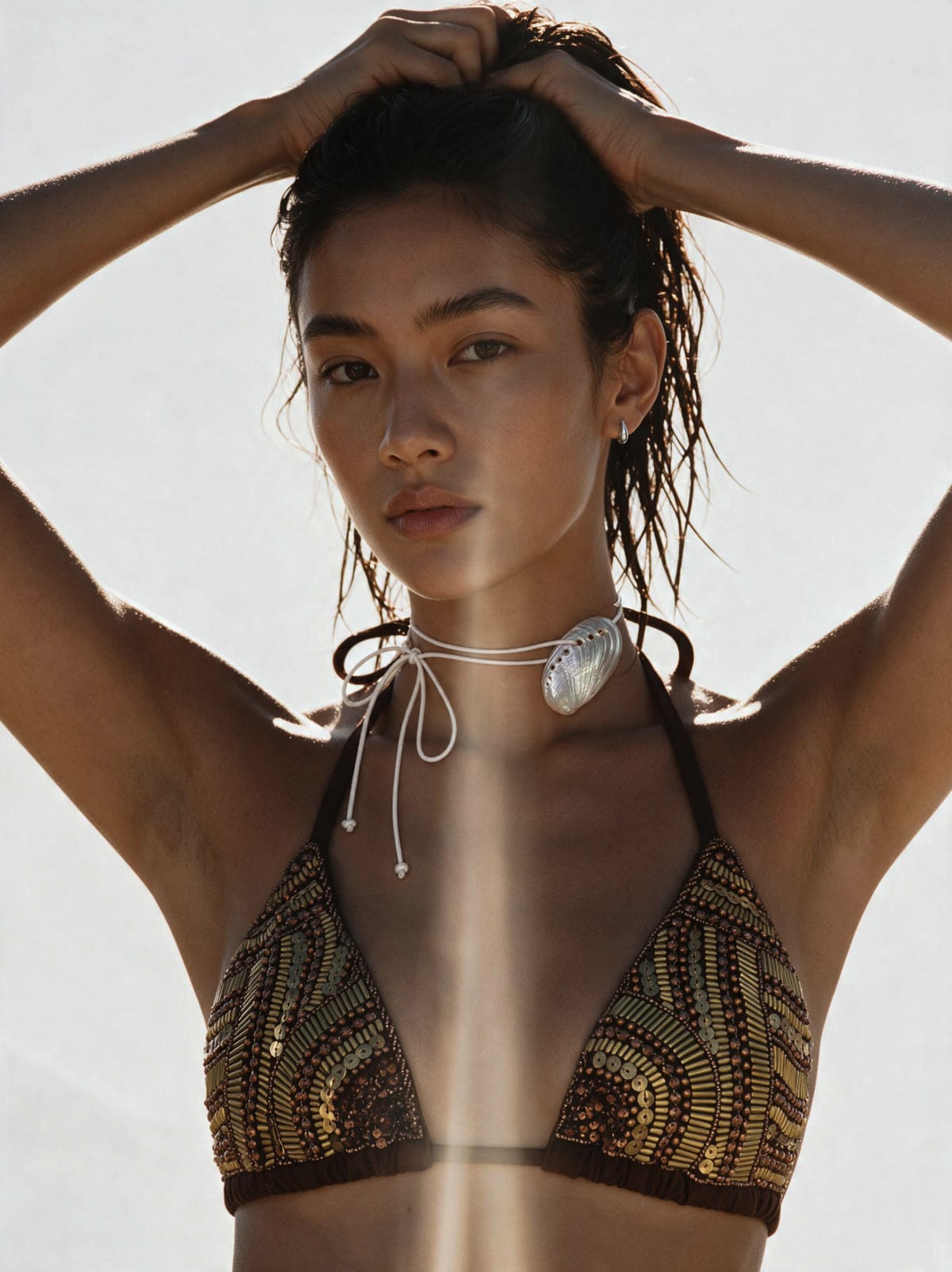 A model with wet hair and a shell pendant holding her hair up, brown bikini with gold beaded triangle cups