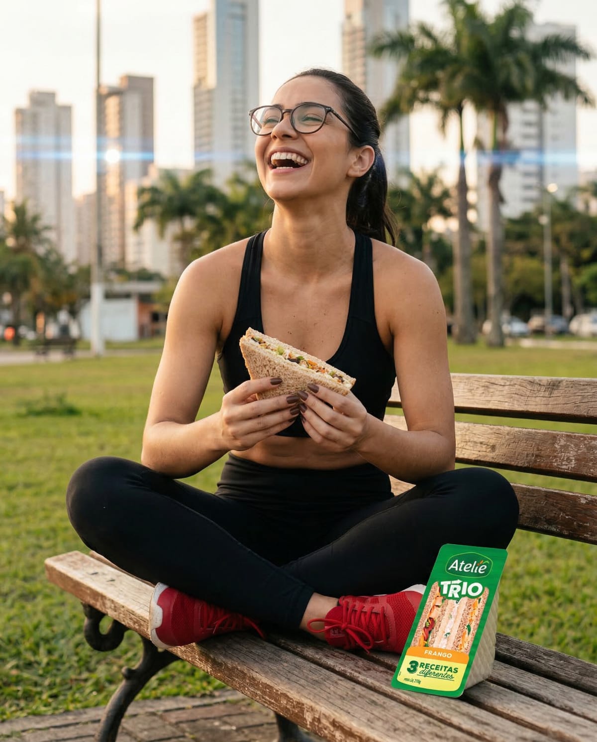 Woman in black sportswear sitting cross legged on a bench with an Ateliê sandwich, city towers behind