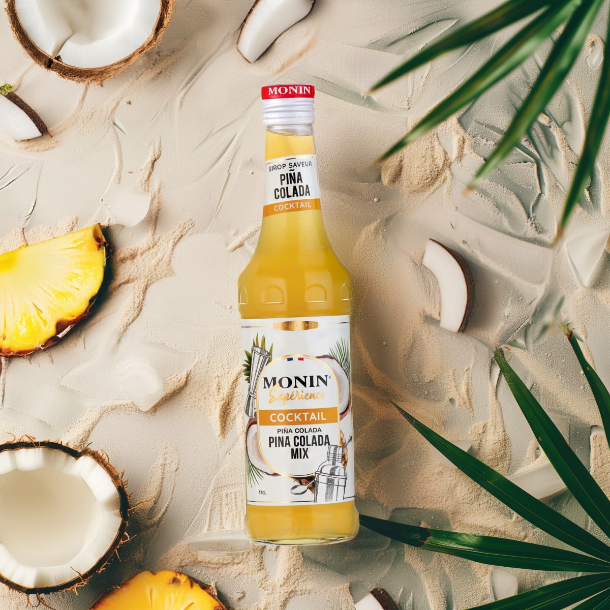 Monin Piña Colada bottle lying on sand with coconut halves, pineapple wedges and palm fronds